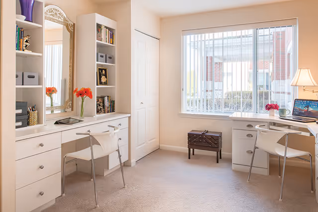 A bright and tidy room with two white desks and chairs positioned on opposite walls. One desk has built-in shelves with books, storage bins, and decorative items, along with a large mirror and a vase of orange flowers. The other desk has a laptop, a lamp, a mug, and a small vase with pink flowers. A window with vertical blinds lets in natural light, and a small dark wooden chest sits beneath the window. The room has beige walls and carpeted flooring.