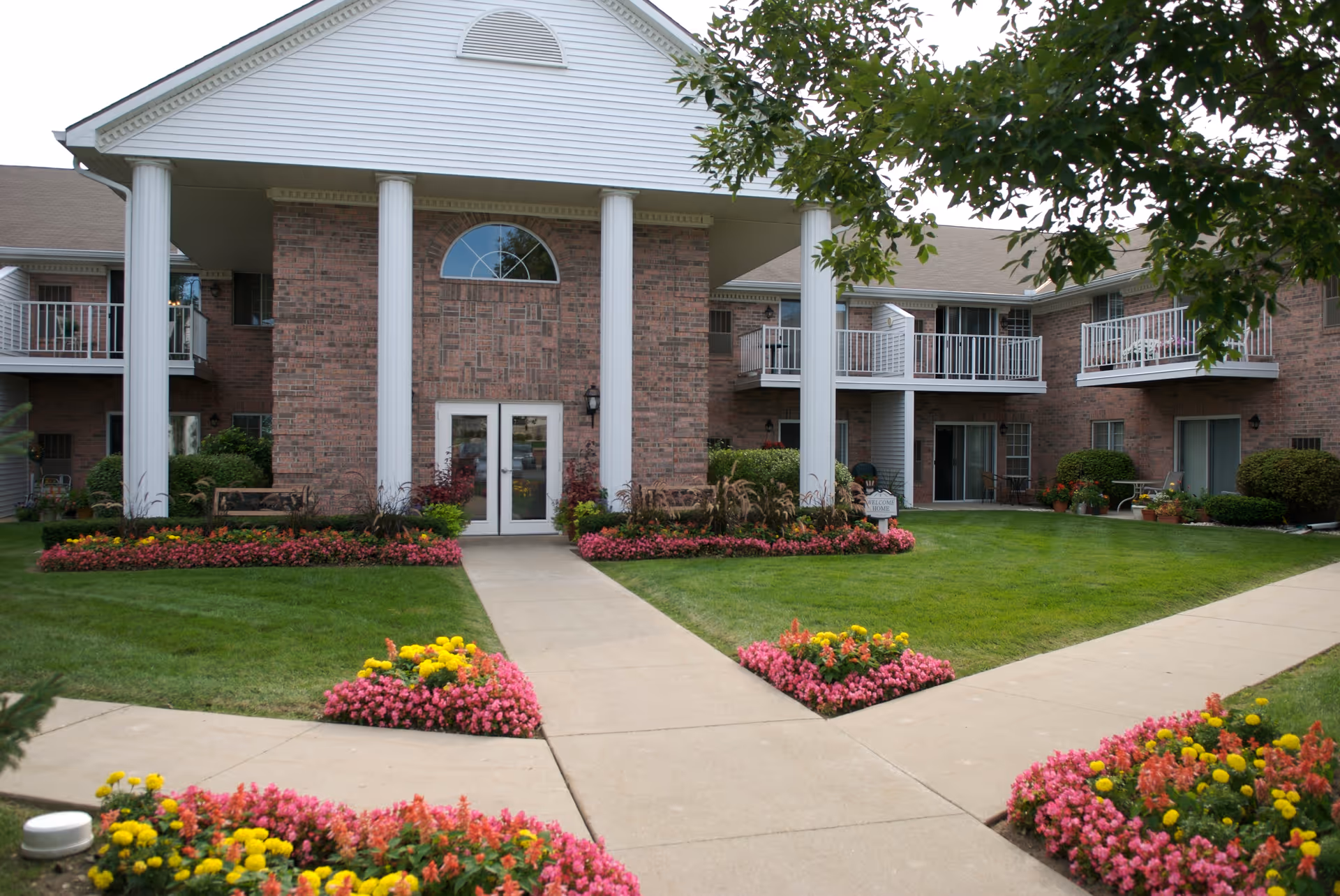 Exterior view of a brick apartment building with white columns and balconies. The entrance has double glass doors and is surrounded by well-maintained green lawns and colorful flower beds with pink and yellow flowers. Sidewalks lead to the entrance, and there are benches and shrubs near the building.