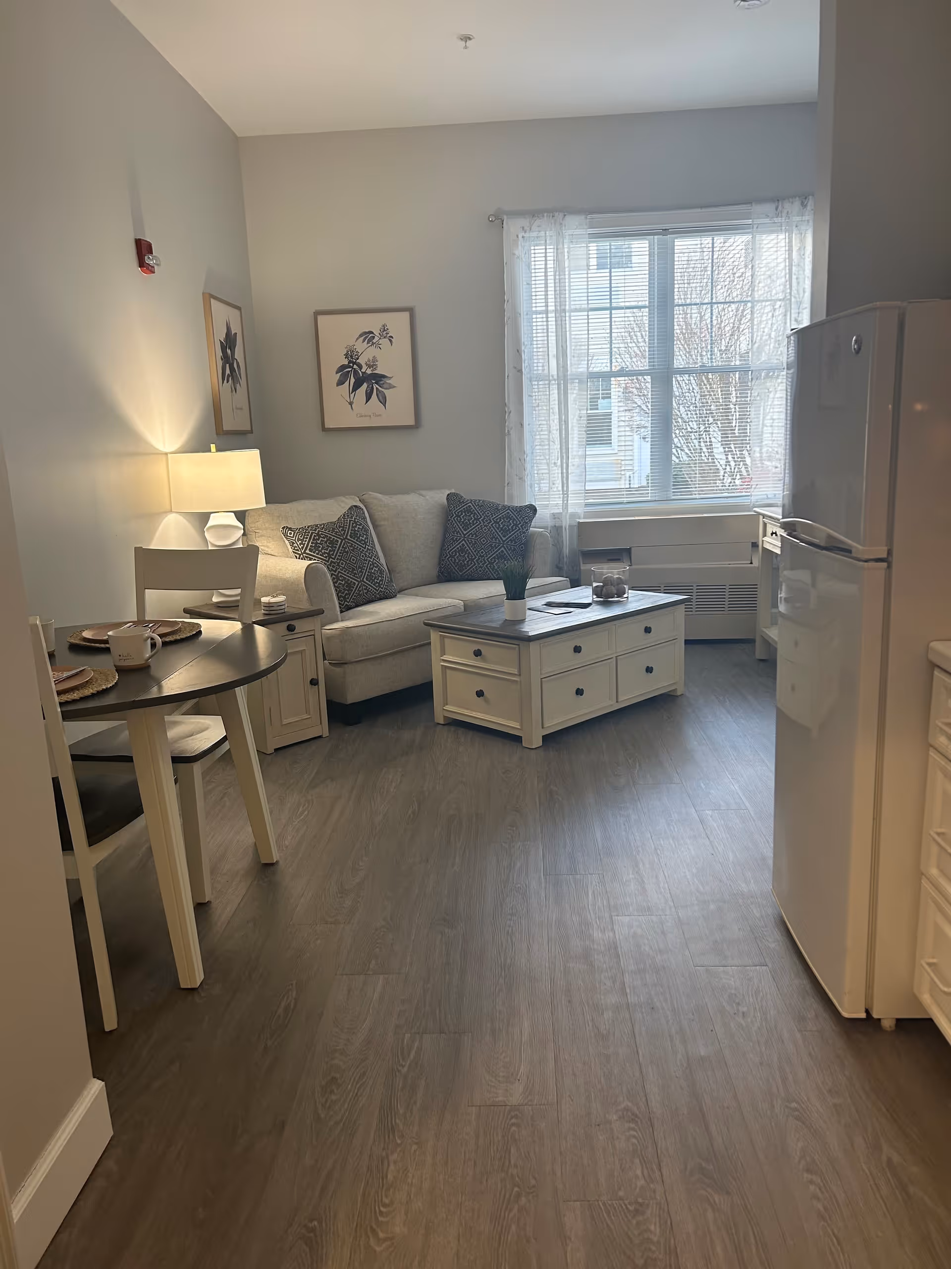 A cozy living area with a beige couch adorned with two patterned pillows, a white coffee table with drawers, and a small dining table set with two chairs and place settings. The room has light gray walls, wood flooring, a window with sheer white curtains, and a white refrigerator partially visible on the right side.
