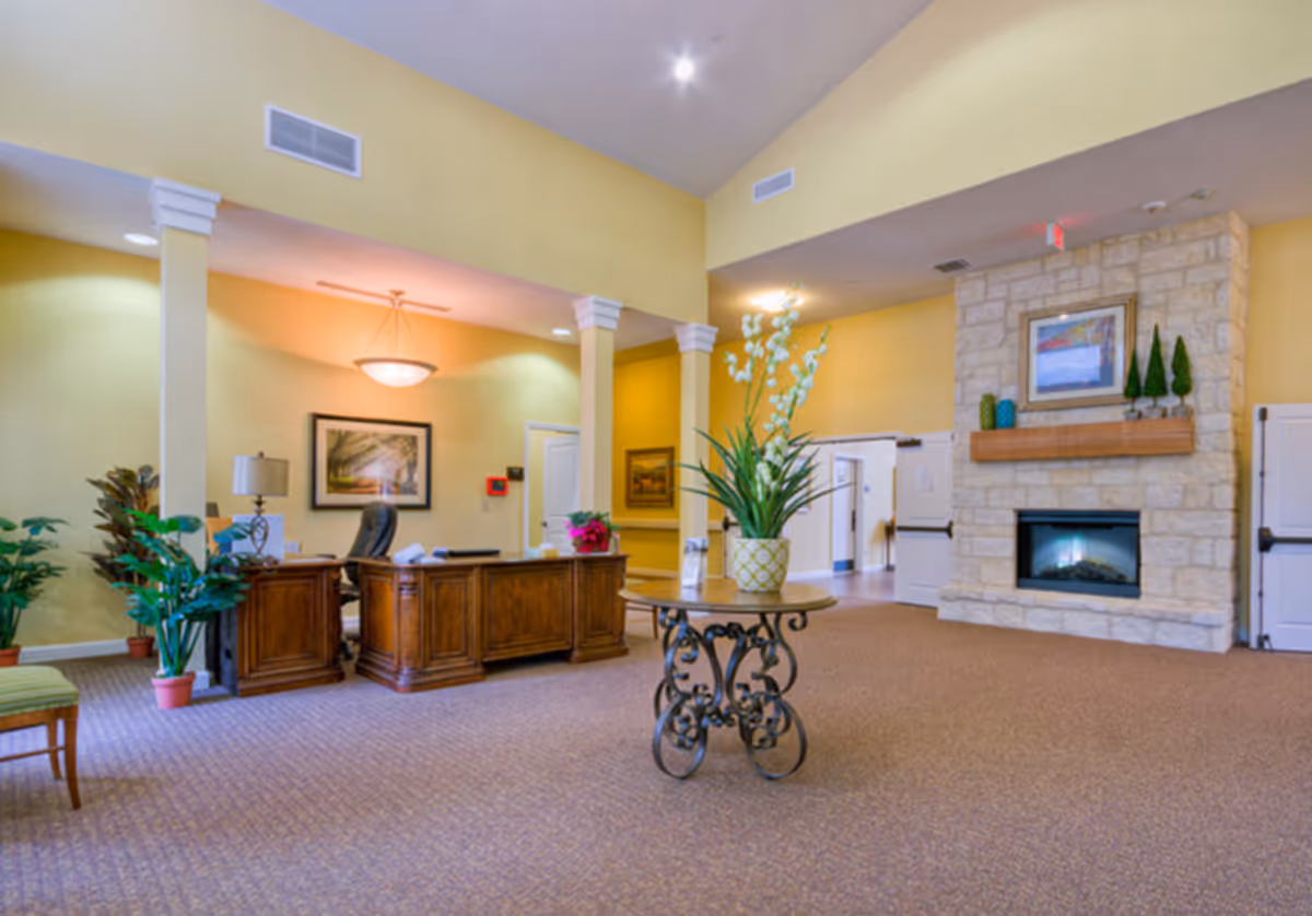 A spacious and well-lit interior area with a wooden reception desk, a round table with a decorative plant, several potted plants, a stone fireplace with a framed picture above it, and yellow walls with white trim.