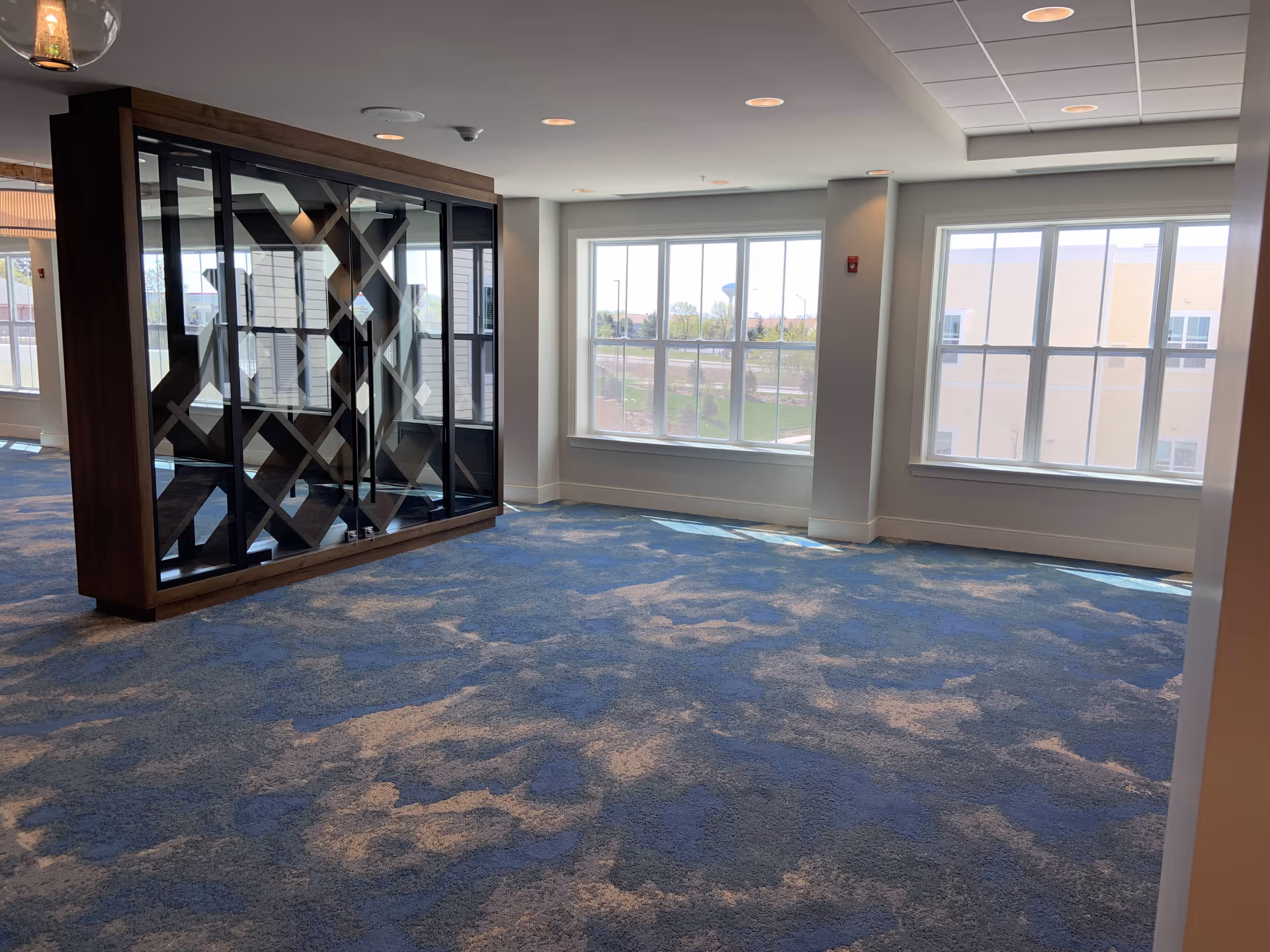 An empty interior room with large windows letting in natural light, blue and beige patterned carpet, and a decorative wooden and glass wine rack or display case. The ceiling has recessed lighting and a drop ceiling section.
