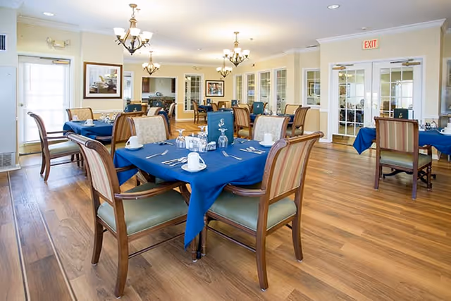 A bright dining room with wooden floors and multiple tables covered with blue tablecloths. Each table is set with white cups, plates, silverware, and glassware. The room has beige walls, chandeliers hanging from the ceiling, and large windows letting in natural light. There are wooden chairs with cushioned seats around the tables.