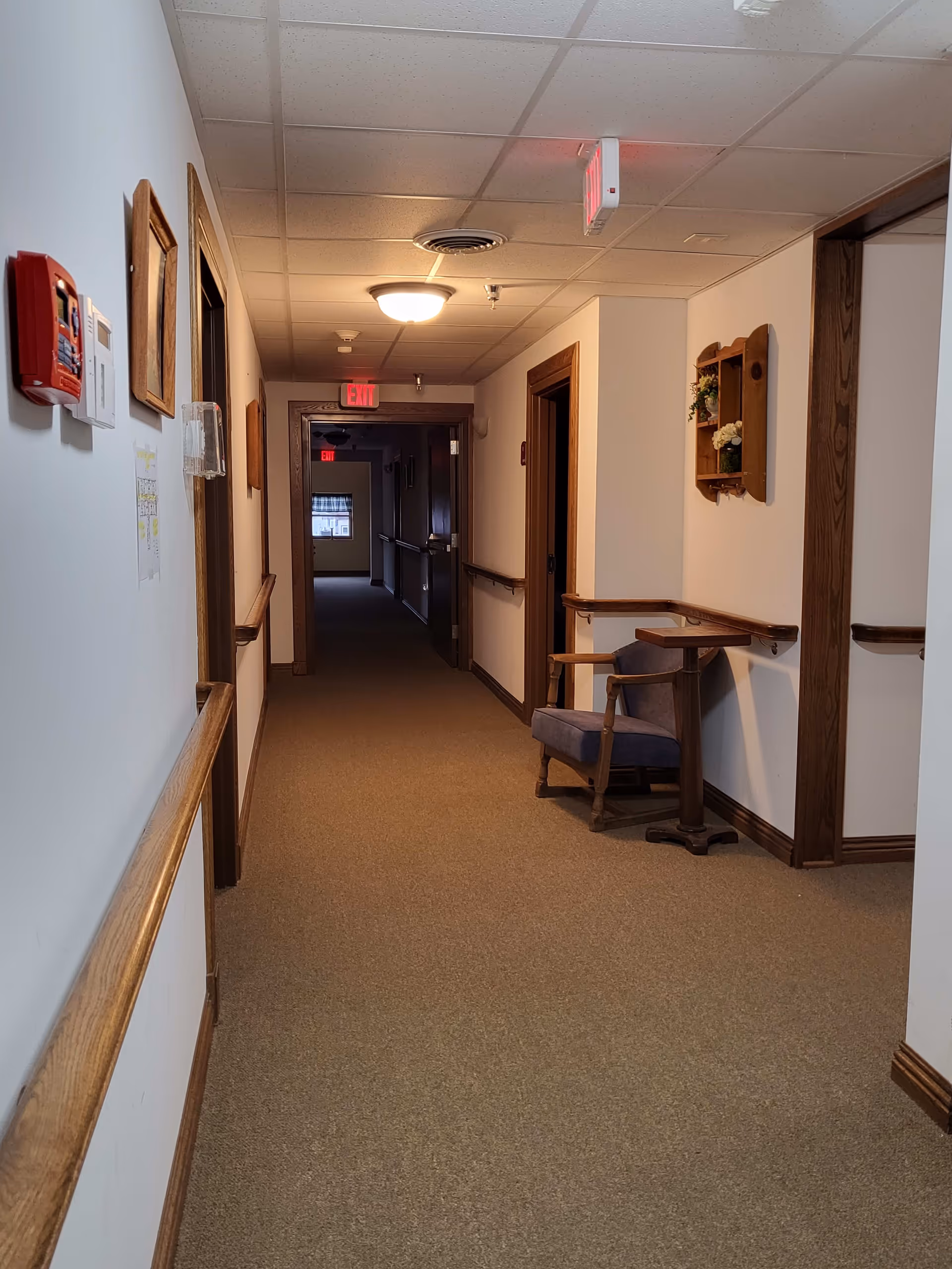 A carpeted hallway in a senior living facility with white walls and wooden trim. There are handrails on both sides of the hallway, a blue upholstered chair with wooden arms and a small attached table on the right side. The hallway is lit by ceiling lights and has exit signs visible at the far end. There are framed pictures and a small wooden shelf with flowers on the walls.