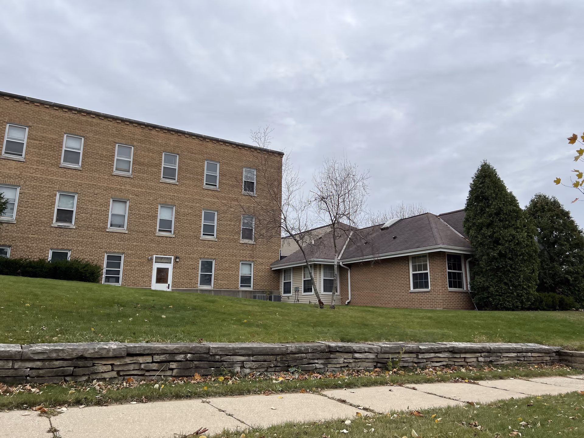 Exterior view of a senior living facility showing a multi-story brick building on the left and a single-story brick building on the right, with a grassy lawn and a stone retaining wall in the foreground under a cloudy sky.