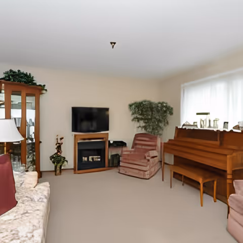 A cozy living room with a beige carpet, a floral patterned sofa with a red pillow, a wooden piano with a bench, a striped armchair, a flat-screen TV mounted above a wooden fireplace, a tall green plant, and a glass-front wooden cabinet with decorative items.