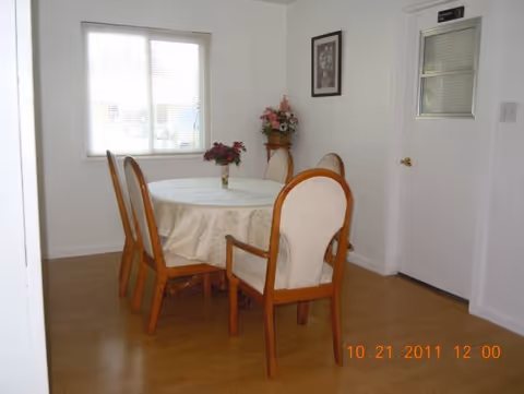 A small dining area with a round table covered with a white tablecloth and four wooden chairs with cushioned seats. A vase with flowers is placed on the table. There is a window with blinds on the left wall and a framed picture hanging on the wall above a small stand with a flower arrangement. A white door with a small window and a sign is on the right side. The floor is wooden.