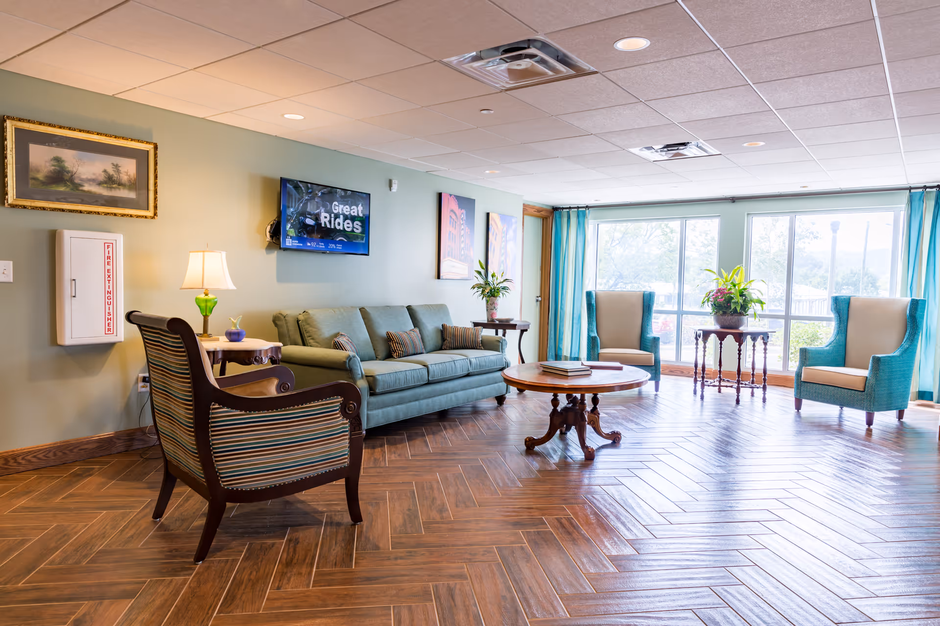 Bright sitting area with a sofa, several armchairs, a round coffee table and large windows in a senior living lounge.