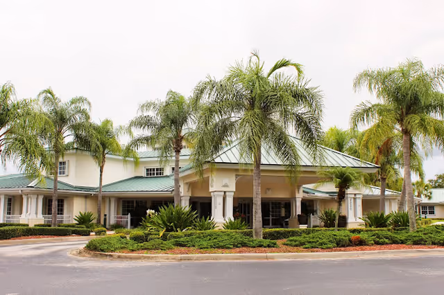 Front entrance of a single-story senior living facility with a covered porte-cochère, palm trees, and landscaped roundabout.