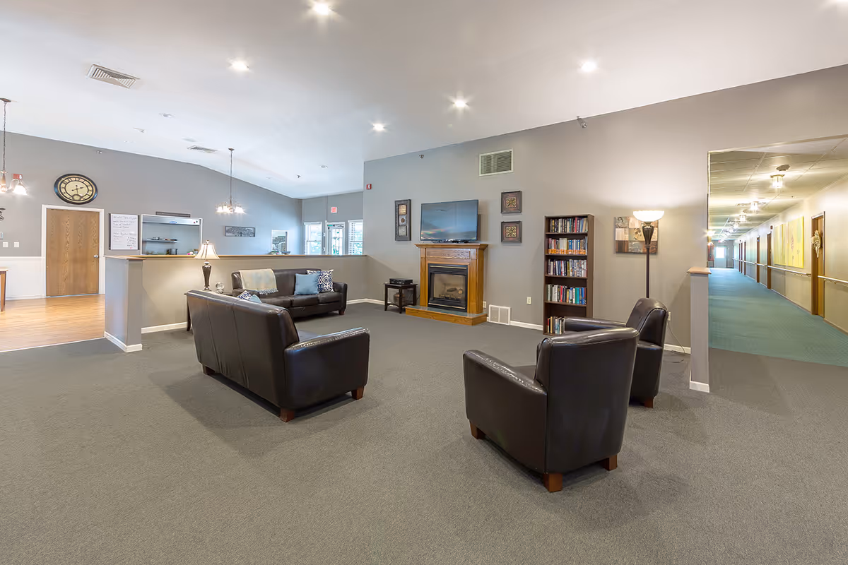 A spacious senior living facility common area with two brown leather sofas and two matching armchairs arranged around a wooden fireplace with a mounted TV above it. There is a bookshelf filled with books next to the fireplace and a floor lamp beside one of the armchairs. The room has gray walls, recessed ceiling lights, and a gray carpet. To the left, there is a partial view of a dining or kitchen area with wooden flooring and a clock on the wall. To the right, a hallway with green carpet and handrails leads to other rooms.