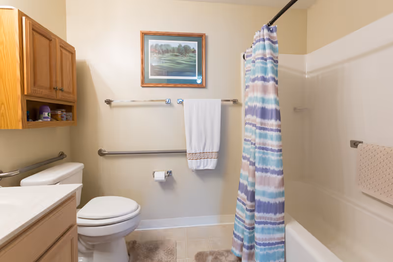 A clean bathroom with a toilet, wooden cabinet, white countertop, and a bathtub with a colorful striped shower curtain. There are two towel bars on the wall, one holding a white towel and the other empty. A framed picture hangs above the towel bars. The floor has beige tiles and two beige bath mats.