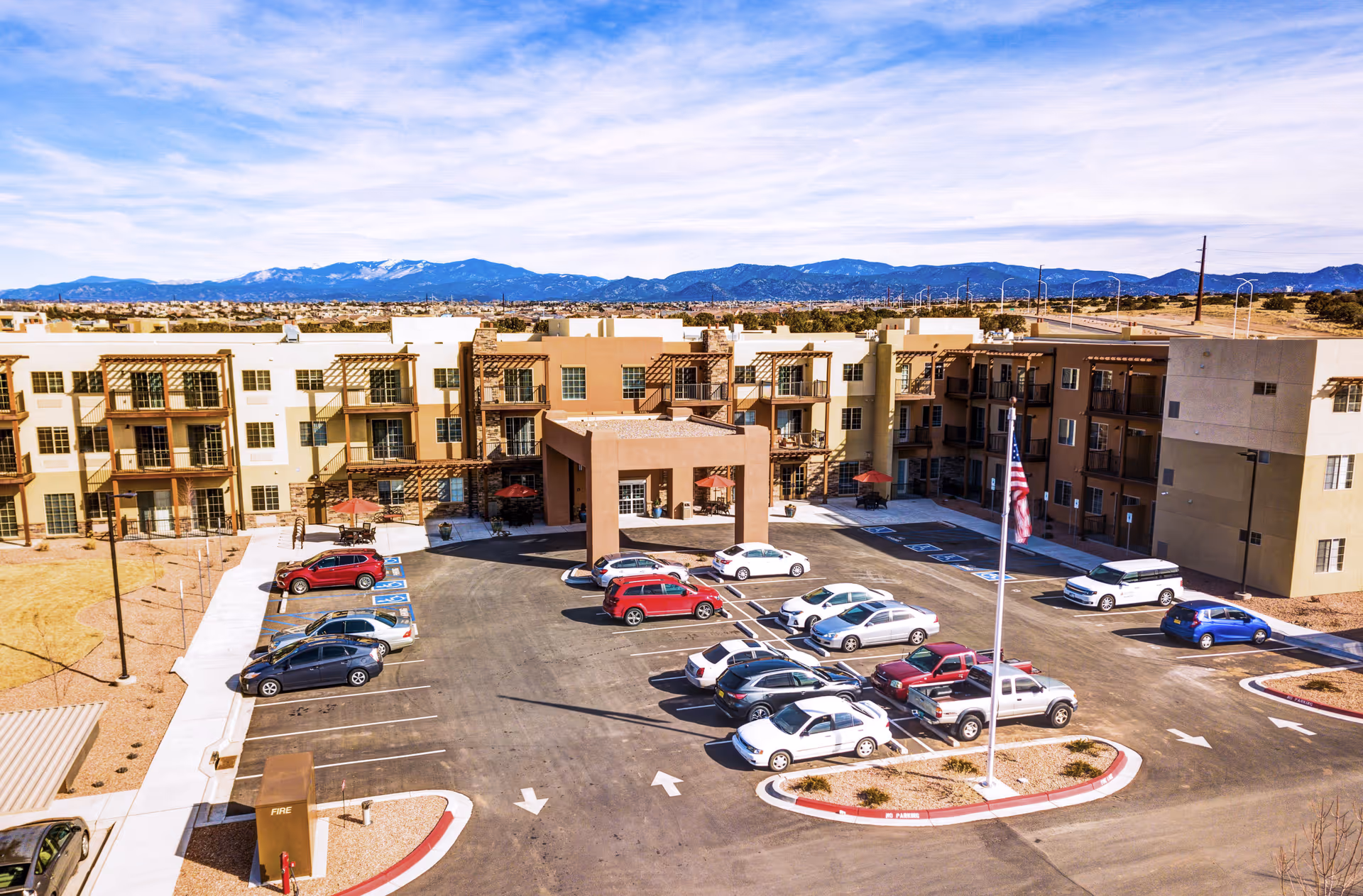 Exterior view of Las Soleras Senior Living facility showing a multi-story building with balconies, a parking lot with several cars, an American flag on a flagpole, and mountains in the background under a partly cloudy sky.
