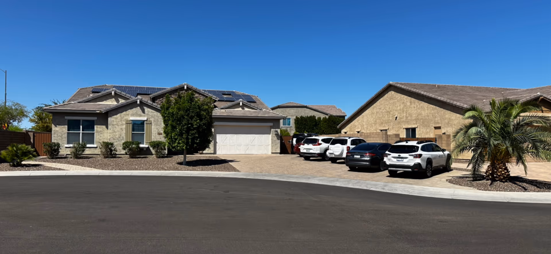 Exterior view of a residential-style building with a garage, several parked cars, and desert landscaping including palm trees and shrubs under a clear blue sky.