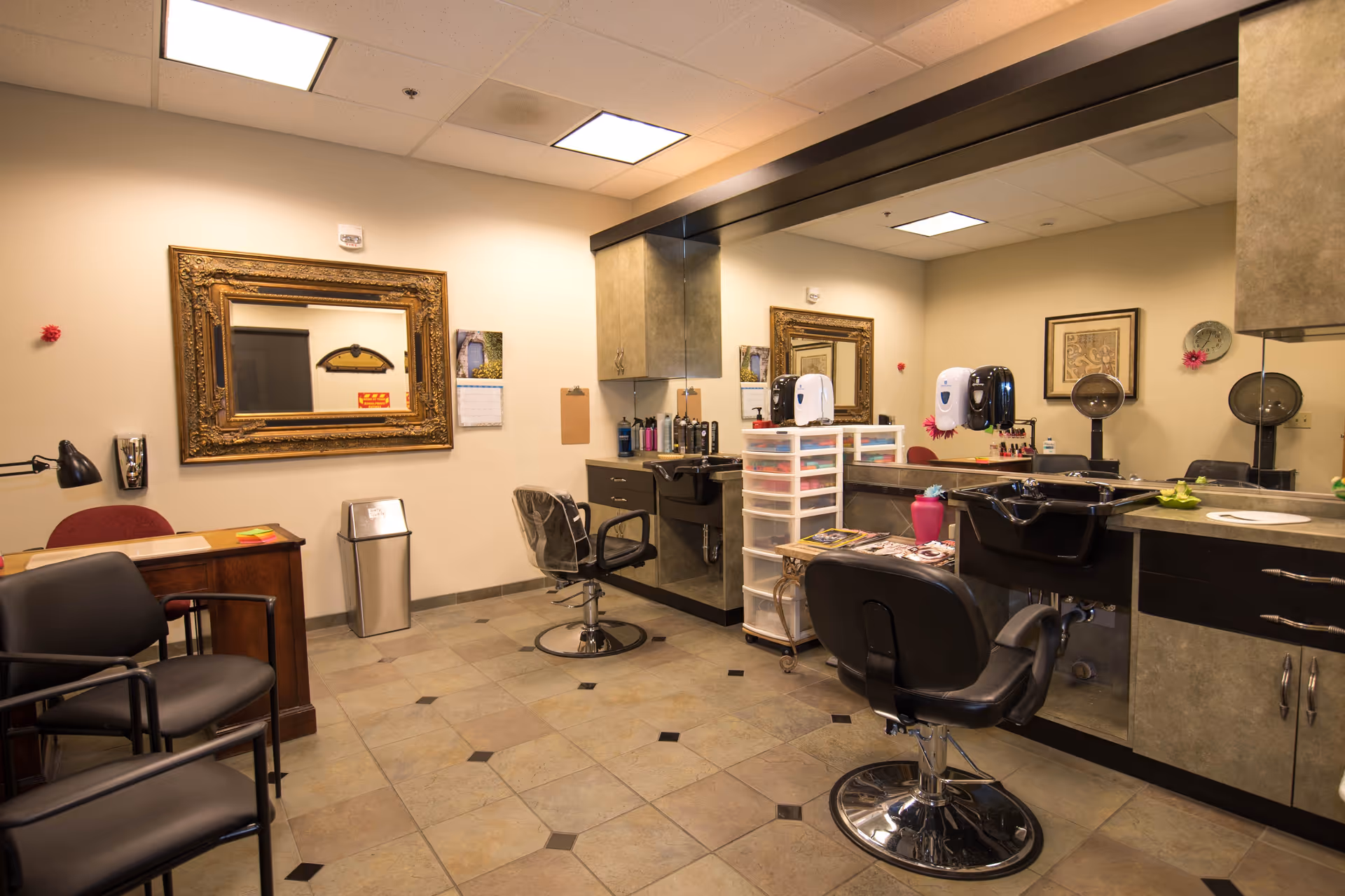 Interior view of a hair salon area within a senior living facility, featuring two salon chairs in front of large mirrors, a sink for washing hair, a small desk with a lamp, and various hair care products and magazines on a plastic drawer unit. The room has tiled flooring, beige walls, and ceiling lights.