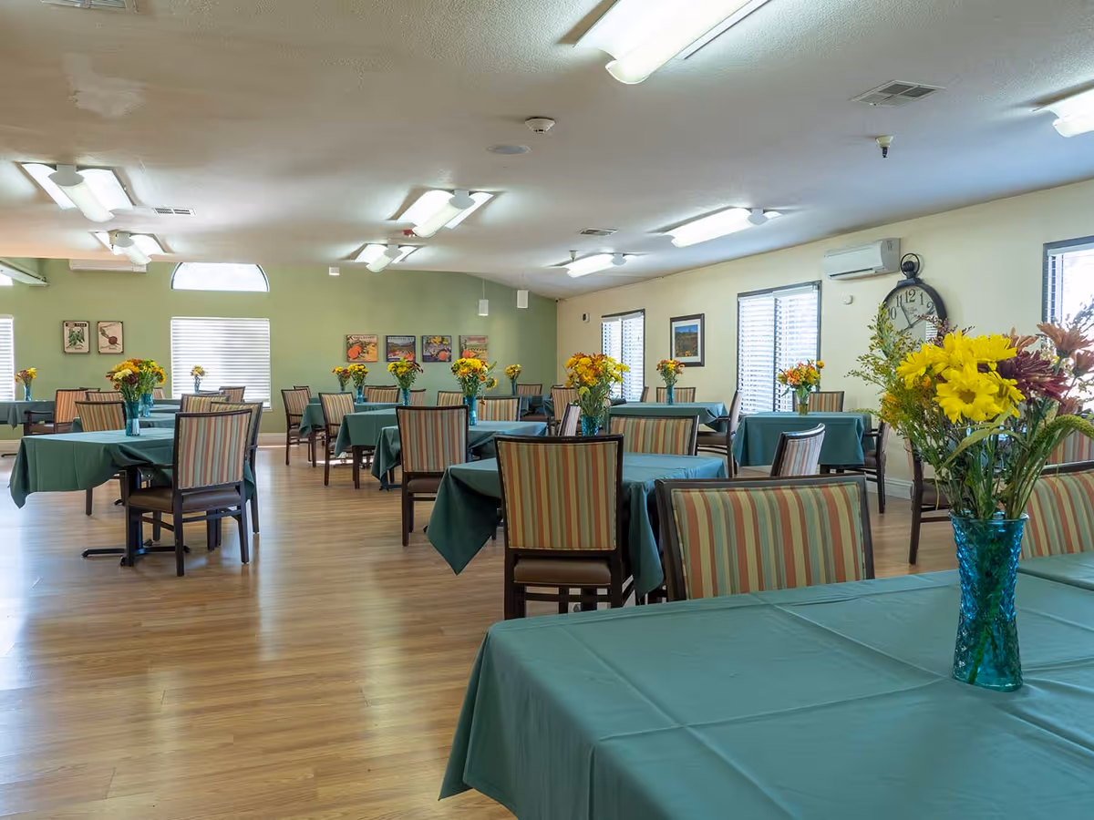 A spacious dining room with multiple tables covered in green tablecloths, each adorned with a vase of colorful flowers. The room has wooden flooring, striped cushioned chairs, several windows with blinds, framed pictures on the walls, and a large clock mounted on one wall.