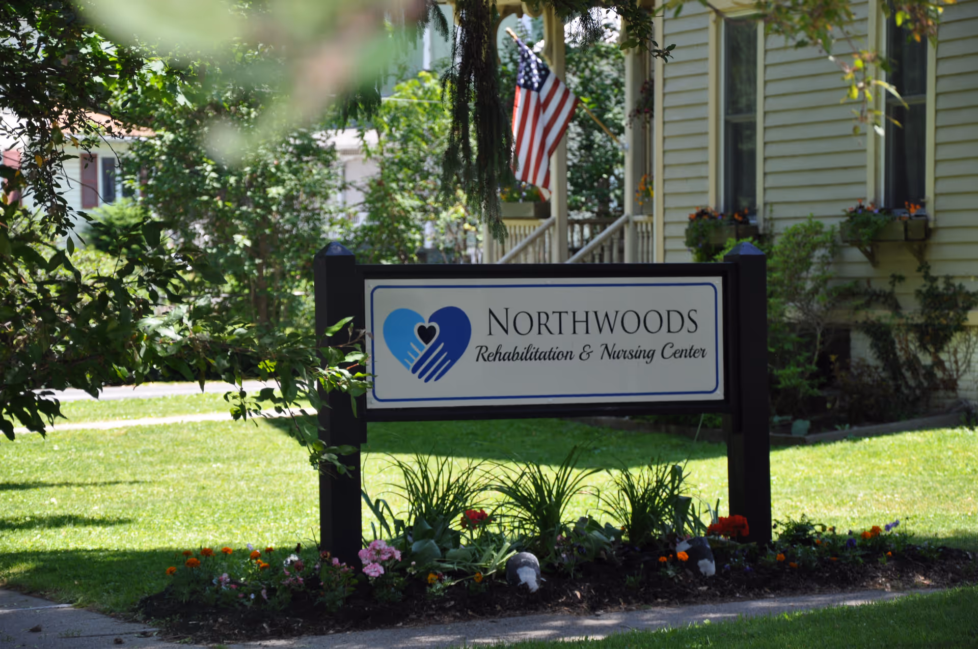 A sign for Northwoods Rehabilitation & Nursing Center is displayed outdoors in a garden area with flowers and greenery. Behind the sign, there is a building with beige siding and windows with flower boxes, and an American flag is visible in the background.