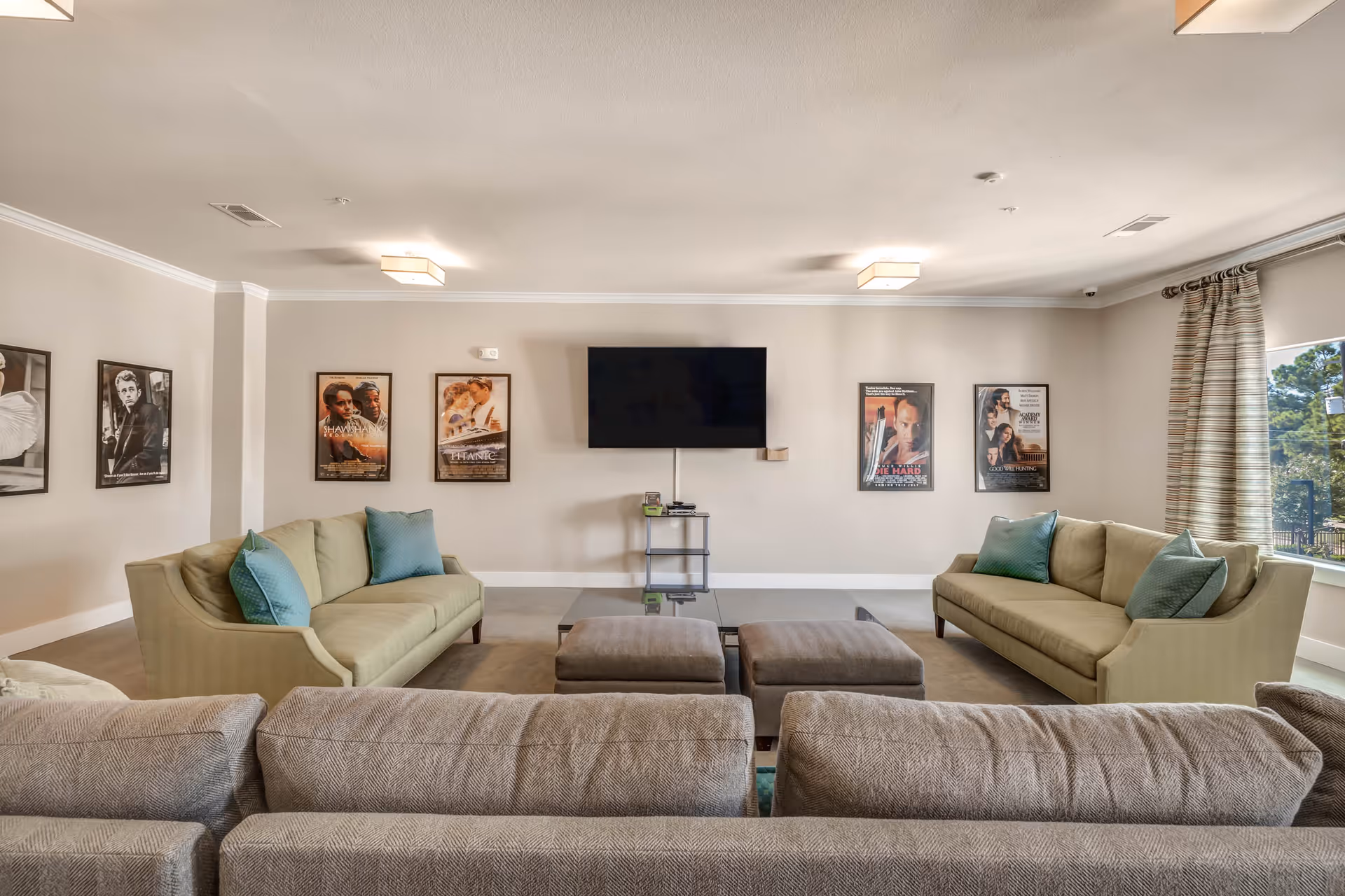 Spacious common lounge with sofas and ottomans facing a wall-mounted TV and framed movie posters.