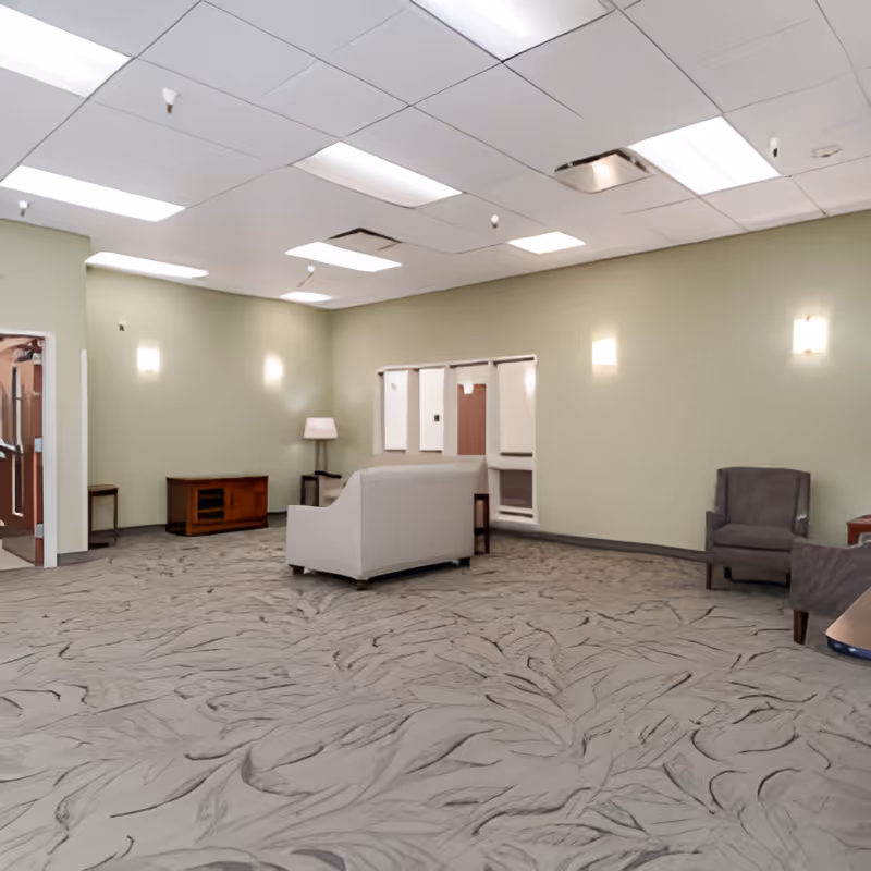 A spacious room with light green walls and a patterned carpet floor. The room contains a white sofa, a gray armchair, a wooden cabinet, a floor lamp, and some small tables. The ceiling has several fluorescent lights and some ceiling tiles are missing or displaced.