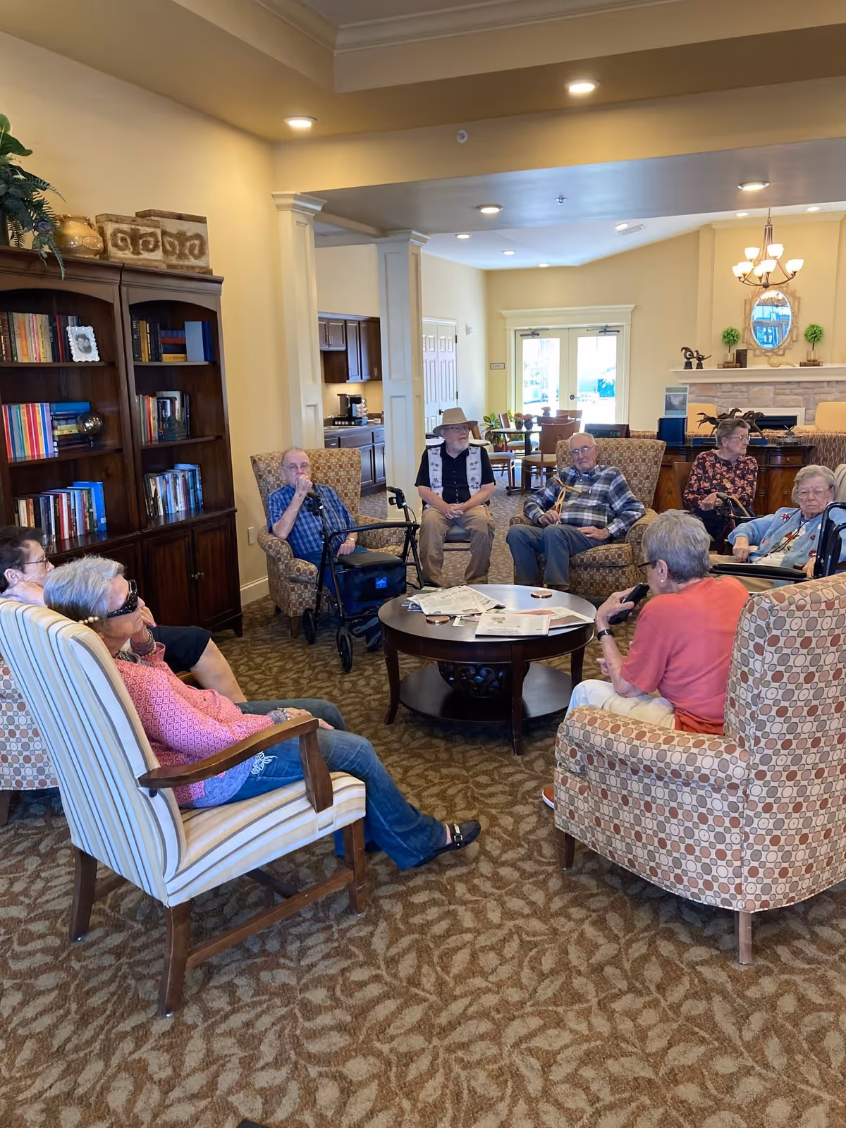 A group of elderly people sitting in a circle on armchairs in a cozy living room area with carpeted floor, a round wooden coffee table with newspapers, bookshelves filled with books, and a fireplace in the background.