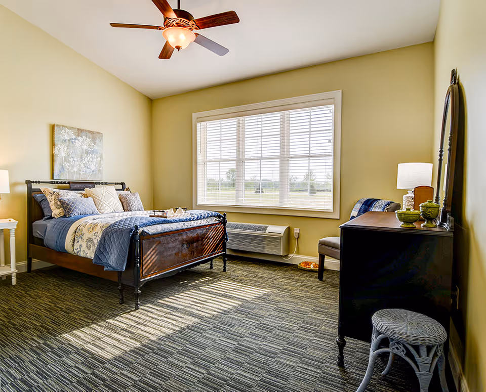 A bright bedroom with a large window letting in natural light. The room features a dark wooden bed with blue and beige bedding, a ceiling fan with a light, a small white side table with a lamp, a dark wooden dresser with a mirror, a chair, and a small wicker stool. The walls are painted light yellow and the floor is carpeted.