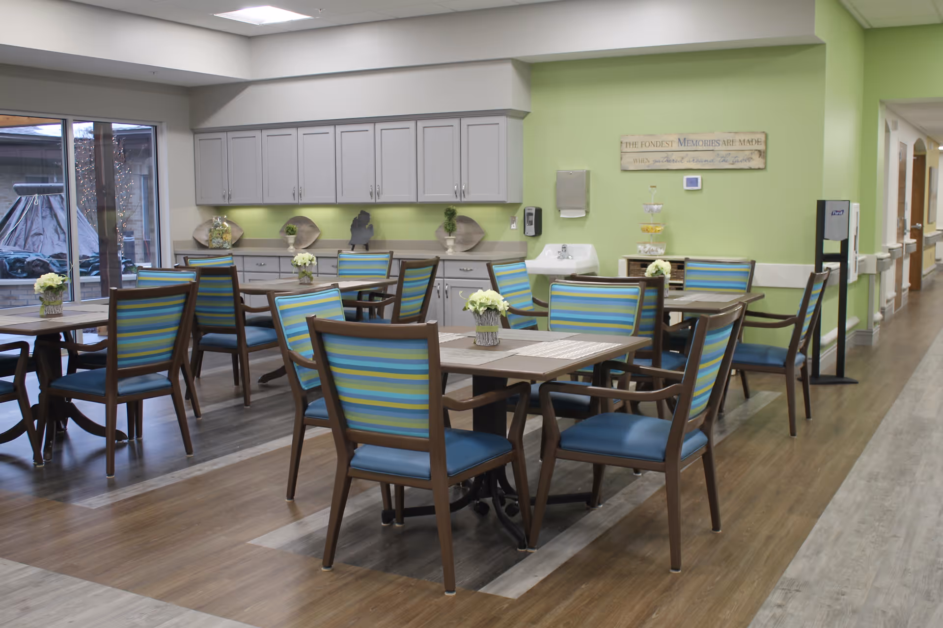 A bright dining area in a nursing and rehabilitation community featuring several wooden tables with blue and green striped cushioned chairs. The room has light green walls, a row of gray cabinets with decorative items on the countertop, a handwashing sink, and a sign on the wall. Large windows provide natural light and a view of an outdoor space.