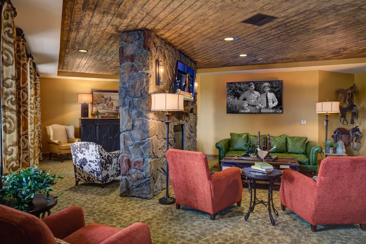 A cozy living room area in a senior living facility featuring a stone fireplace with a mounted TV above it. The room has a green sofa, two red armchairs, and a patterned armchair. There are two floor lamps, a small round table with books, and decorative items including horse sculptures on the wall. The ceiling has a rustic wooden finish and the carpet is patterned.