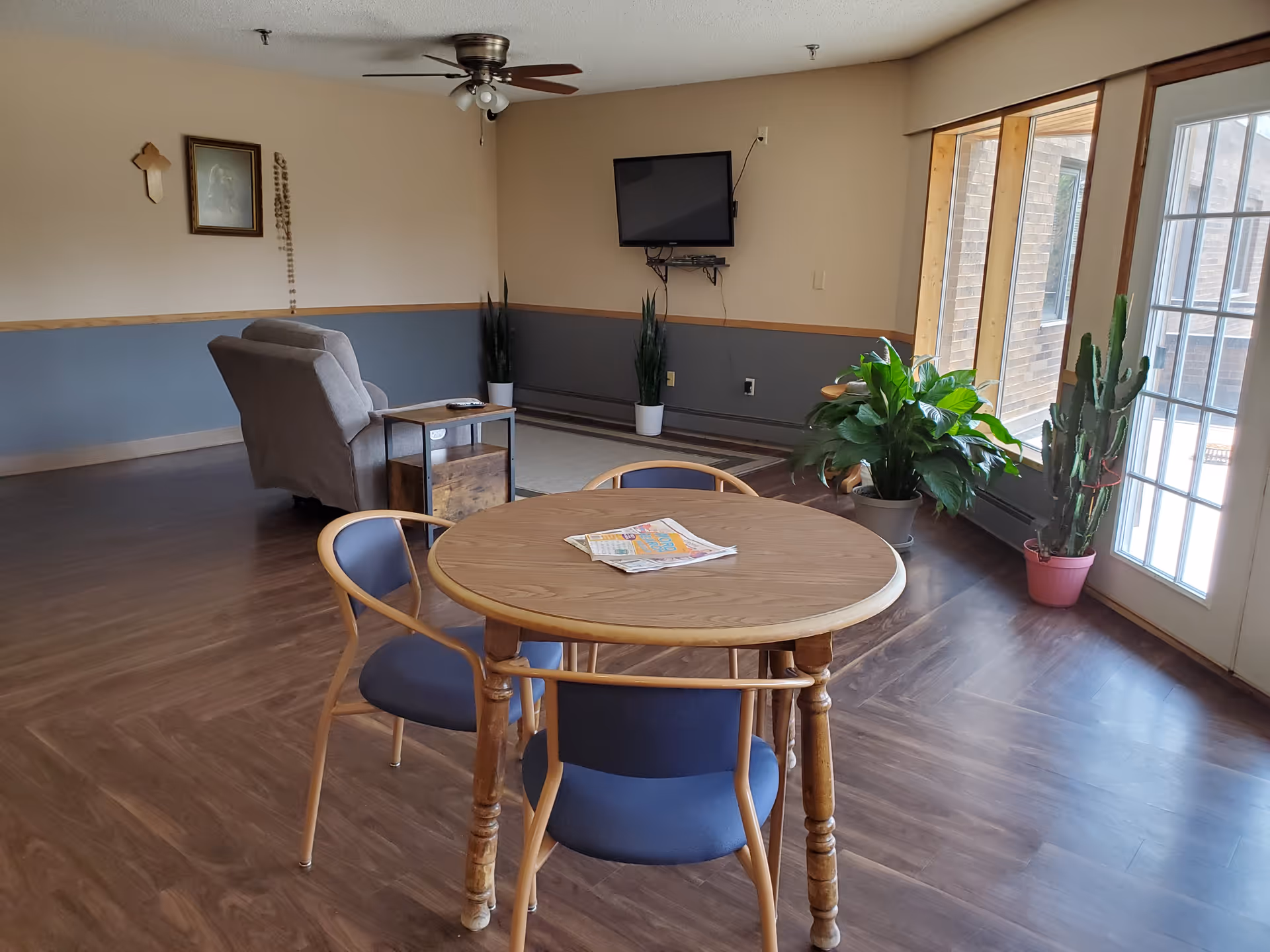 Sunlit common room with a round table and chairs, potted plants, an armchair, wall-mounted TV, and a glass door.
