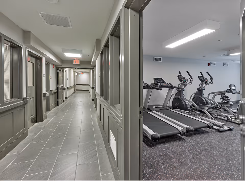 Interior hallway of a senior living facility with a view into a fitness room containing treadmills and elliptical machines.