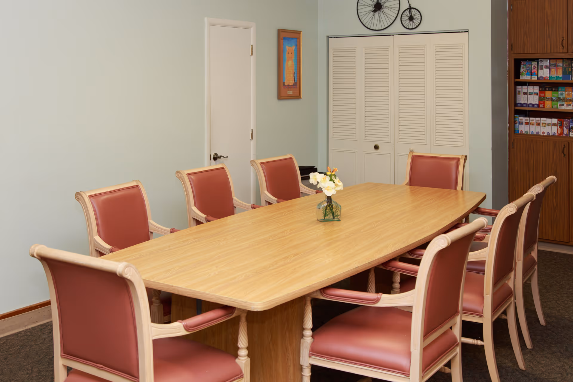 Long wooden dining table surrounded by upholstered chairs in a small dining/meeting room with a vase of flowers on the table.