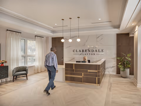 A modern reception area at Clarendale Clayton with a man walking towards the front desk where a receptionist is seated. The space features light wood flooring, a large marble wall with the facility name, three pendant lights hanging from the ceiling, a window with sheer curtains, a small chair, and a potted plant.