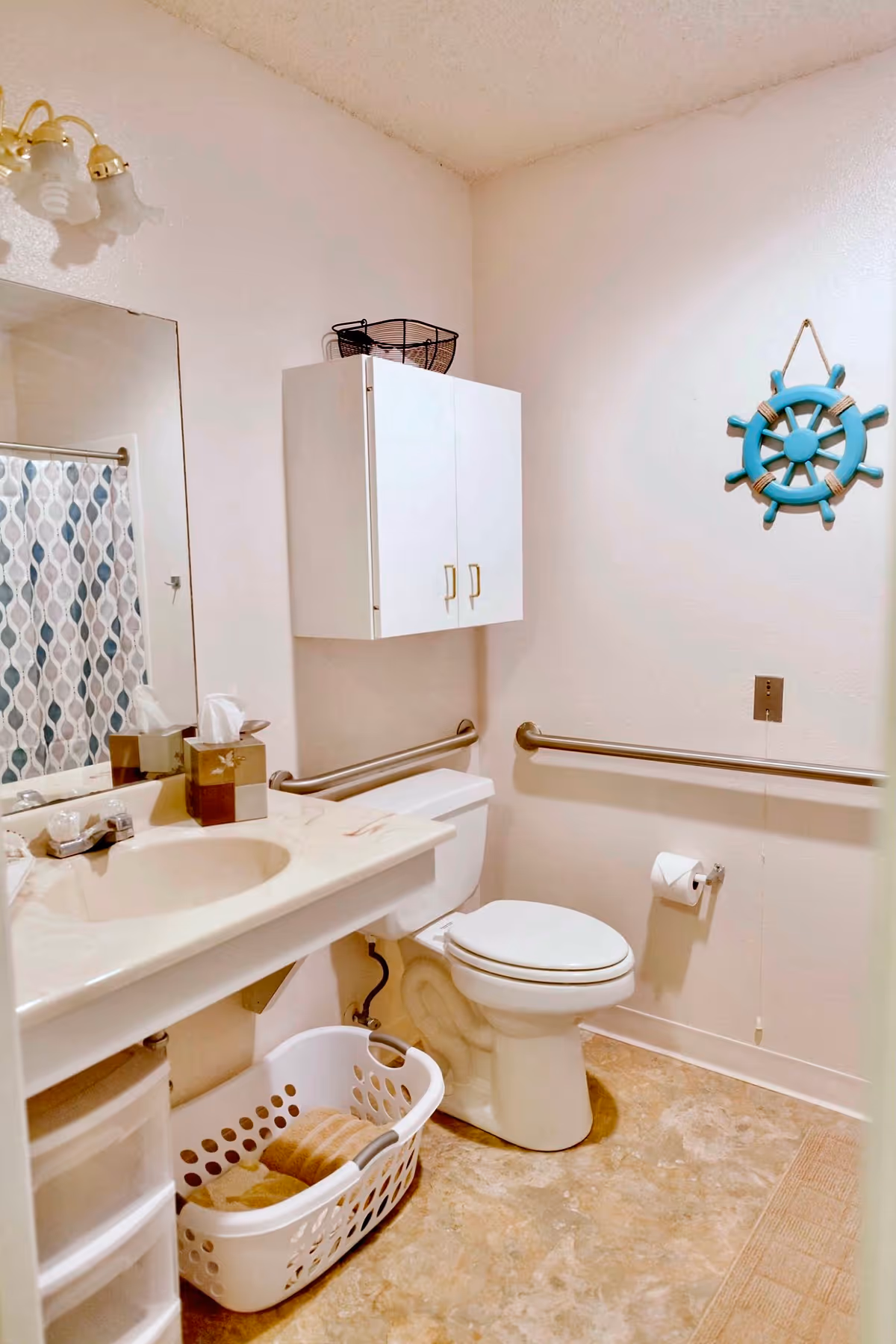 A clean bathroom with a white toilet, a sink with a marble countertop, a large mirror, and a white cabinet mounted on the wall above the toilet. There are grab bars on the walls near the toilet, a laundry basket with towels underneath the sink, a tissue box on the countertop, and a decorative blue ship wheel hanging on the wall.