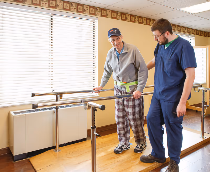 An elderly man wearing a gray jacket, plaid pants, and a baseball cap is using parallel bars for physical therapy while being assisted by a healthcare worker dressed in blue scrubs in a well-lit room with wooden flooring and large windows with blinds.