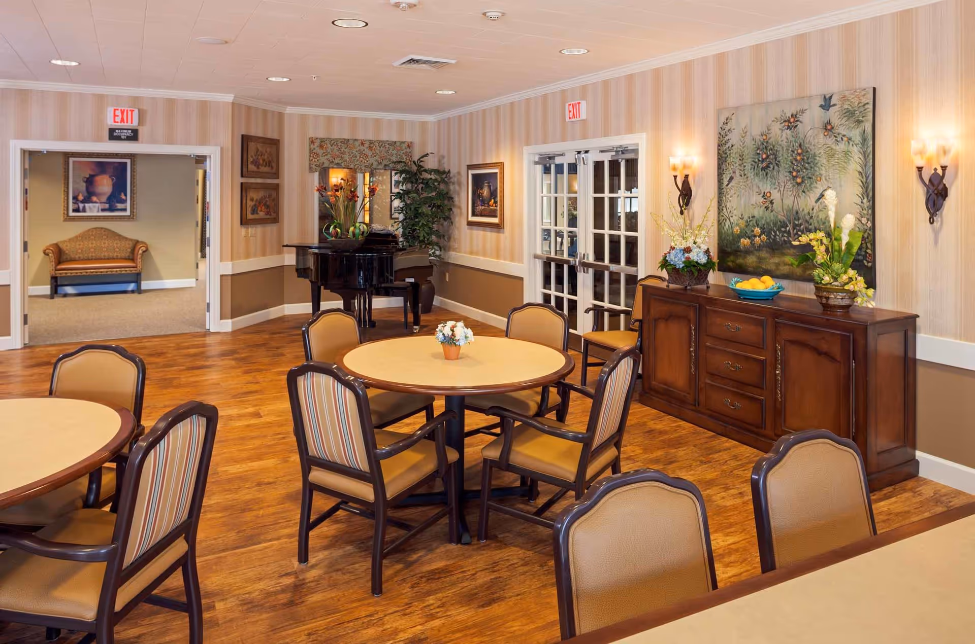 A well-lit room with round tables and chairs arranged for seating, featuring wooden flooring and beige striped wallpaper. There is a wooden sideboard with floral arrangements and a large painting above it. A black grand piano is placed against one wall, and there are exit signs above two doorways, one leading to another room with a patterned loveseat and framed artwork.