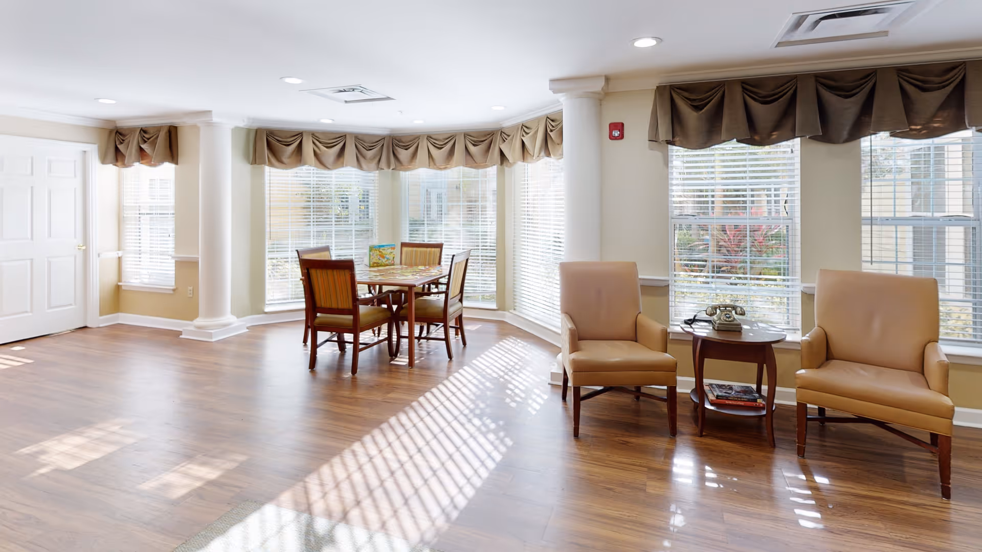 Sunlit common room with wooden floors, a small dining table by bay windows, and two armchairs flanking a side table.