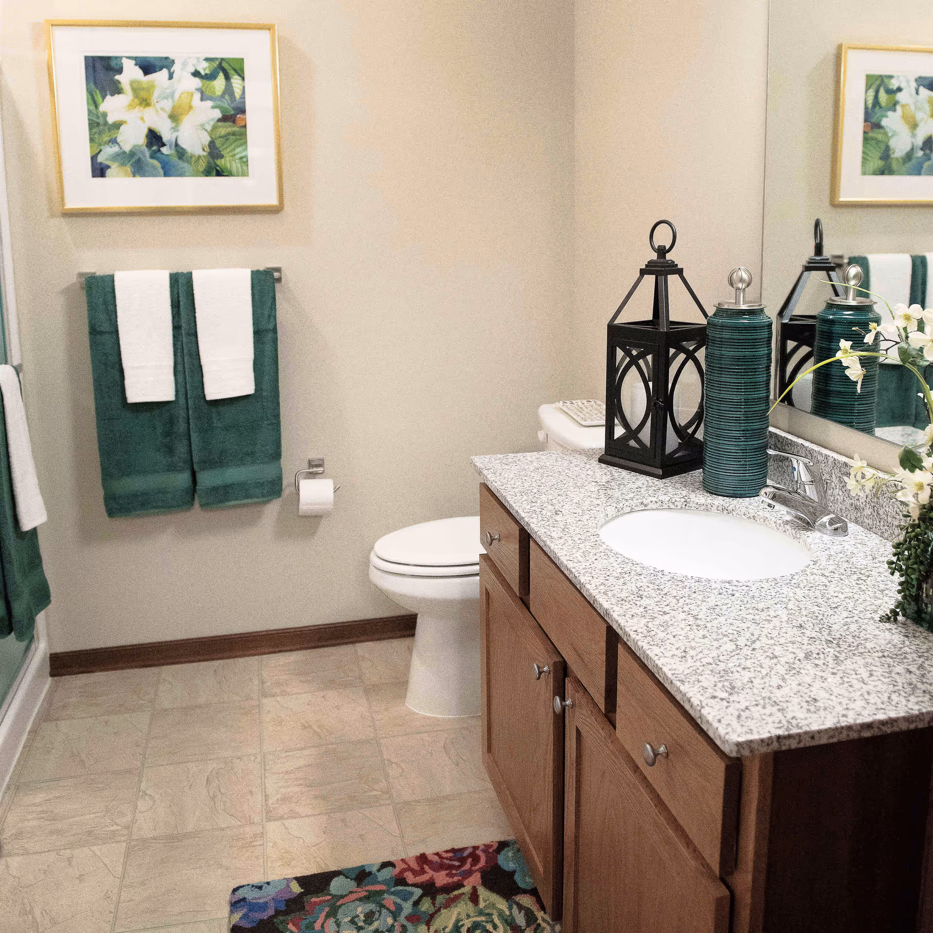 Well-lit bathroom with a granite-topped vanity and sink, toilet, green towels, decorative lanterns, and floral artwork on the wall.