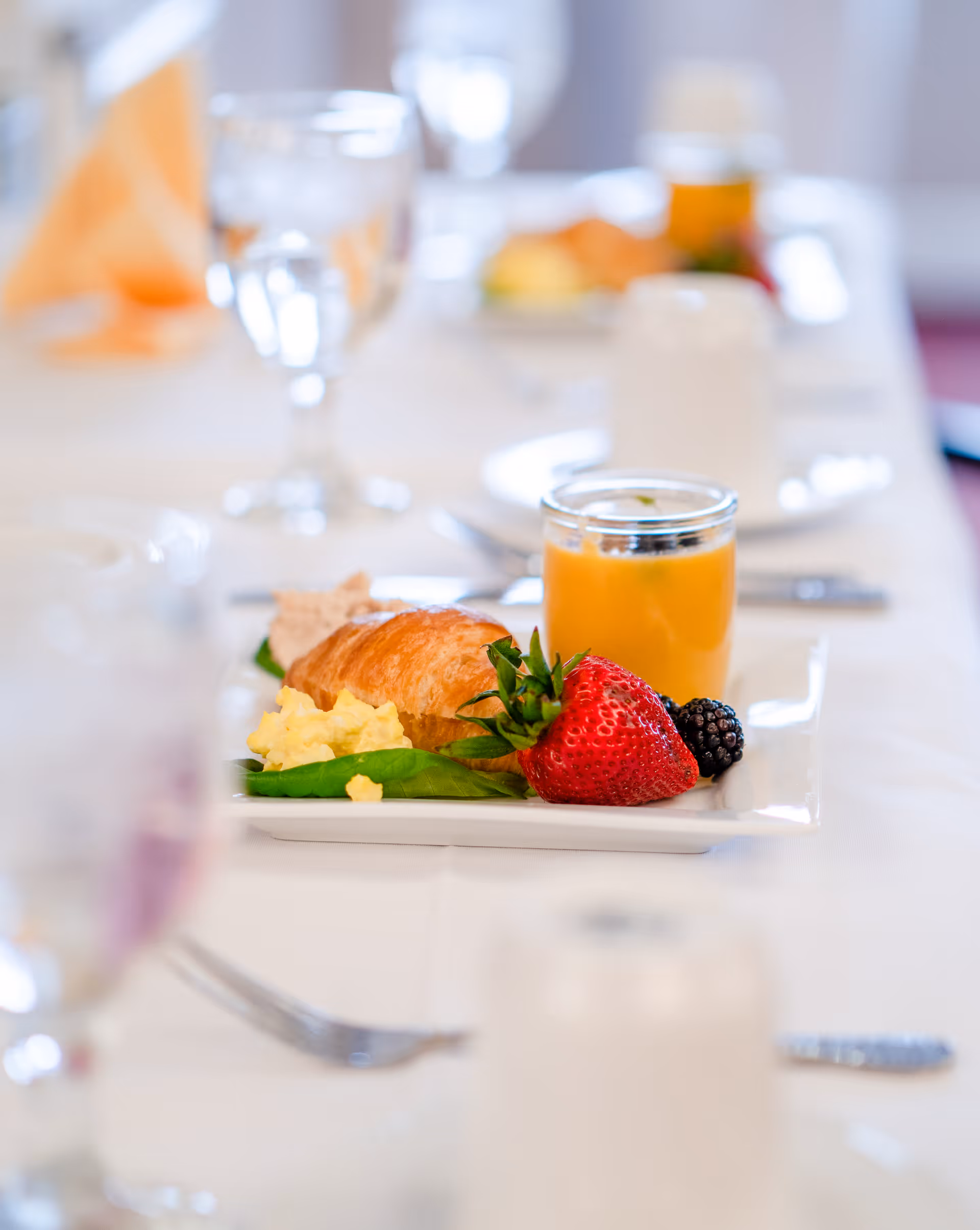 A close-up of a breakfast plate on a white tablecloth featuring a croissant, scrambled eggs on a bed of spinach, a large strawberry, a blackberry, and a small glass jar of orange juice. In the background, there are blurred glasses of water and other plates on the table.