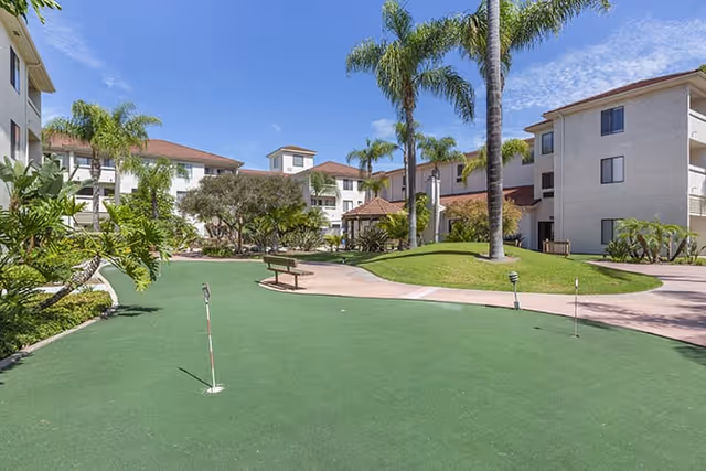 Outdoor putting green area with small golf holes and flags surrounded by palm trees and shrubs, with multi-story residential buildings in the background under a clear blue sky.
