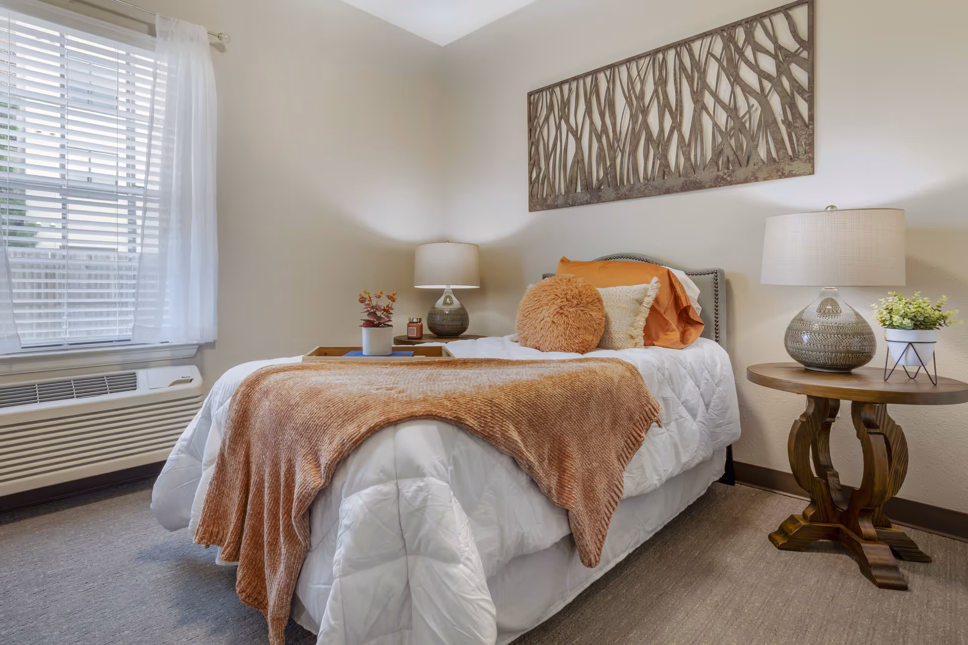 A cozy bedroom with a single bed dressed in white bedding and a soft brown throw blanket. The bed has decorative pillows including a round furry one and is positioned against a beige wall with a metal wall art piece above it. On the right side of the bed is a wooden round side table with a ceramic lamp and a small potted plant. A window with white blinds and sheer curtains is on the left side, and an air conditioning unit is below the window.