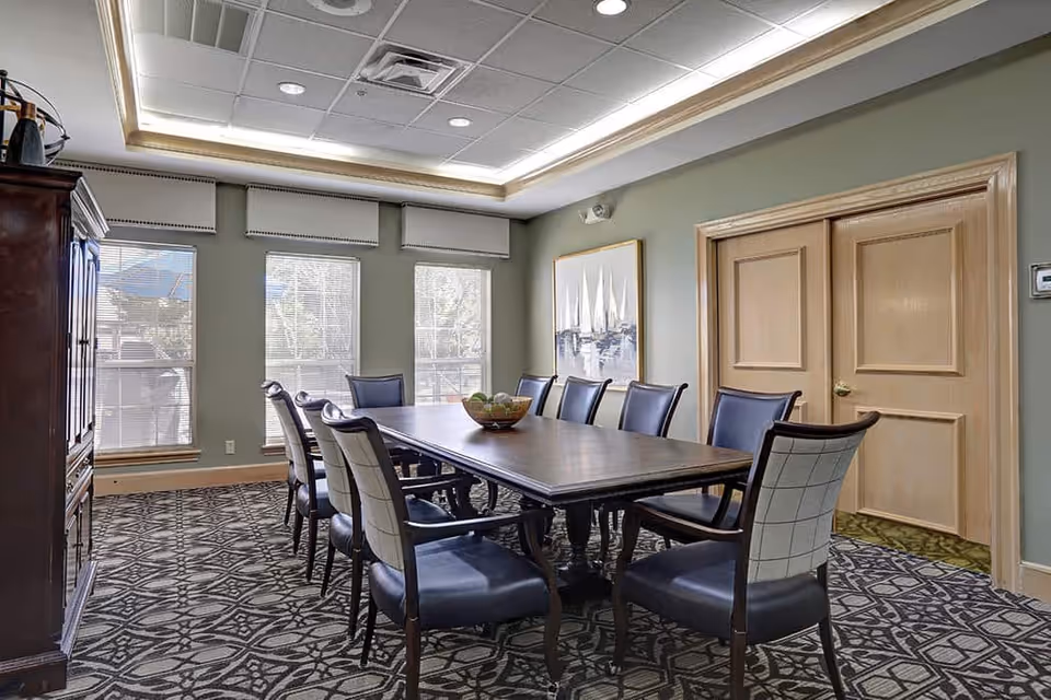 A well-lit dining room with a long wooden table surrounded by ten cushioned chairs. The room has three large windows with blinds, a patterned carpet, light green walls, a wooden cabinet on the left, and a framed painting on the wall. There are double wooden doors on the right side of the room.