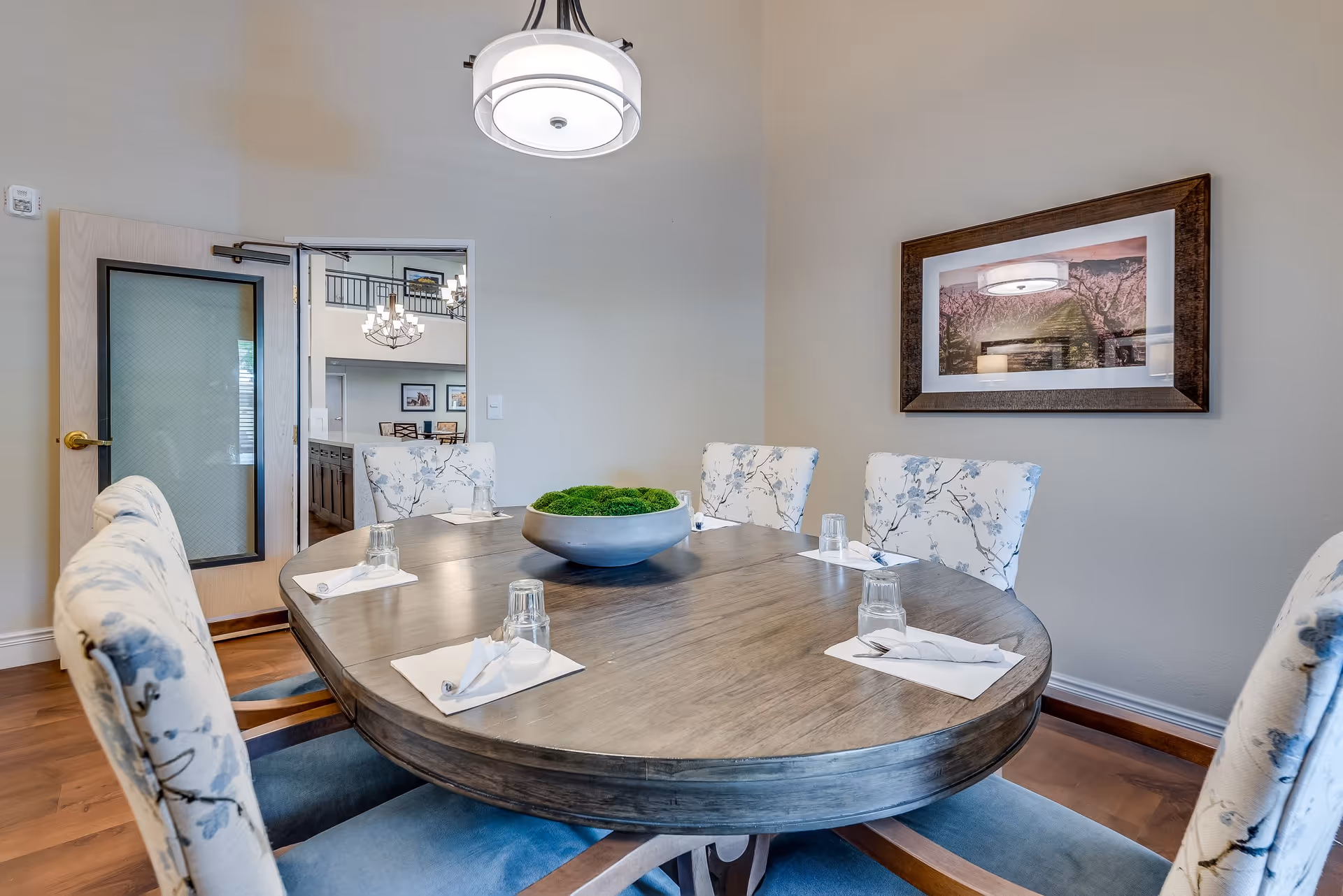 A dining room with a large oval wooden table set with six floral upholstered chairs. Each place setting has a glass, a napkin, and silverware. A decorative bowl with green moss is centered on the table. A modern light fixture hangs above the table, and a framed landscape photograph is on the wall. An open door reveals a view into another room with chandeliers and framed pictures.