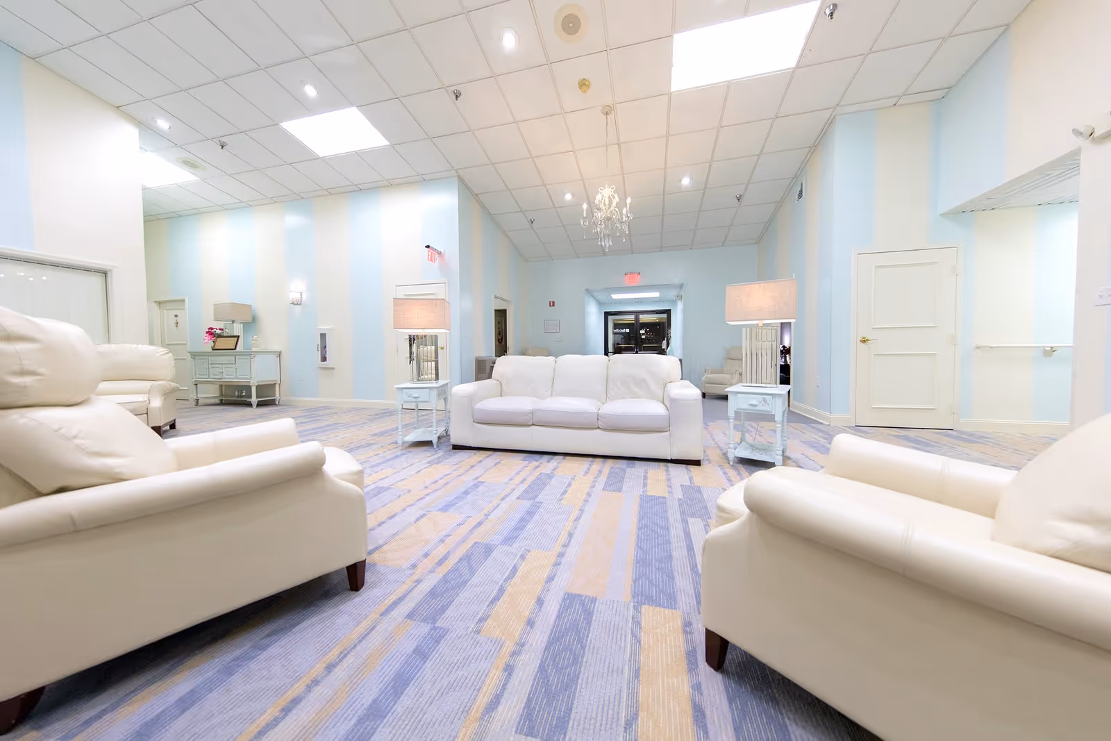 A bright and spacious living room area with white leather sofas and armchairs arranged around two white side tables with lamps. The room has light blue and white striped walls, a patterned carpet in shades of blue and beige, and a chandelier hanging from the ceiling. There is a doorway at the back and additional seating visible in the background.