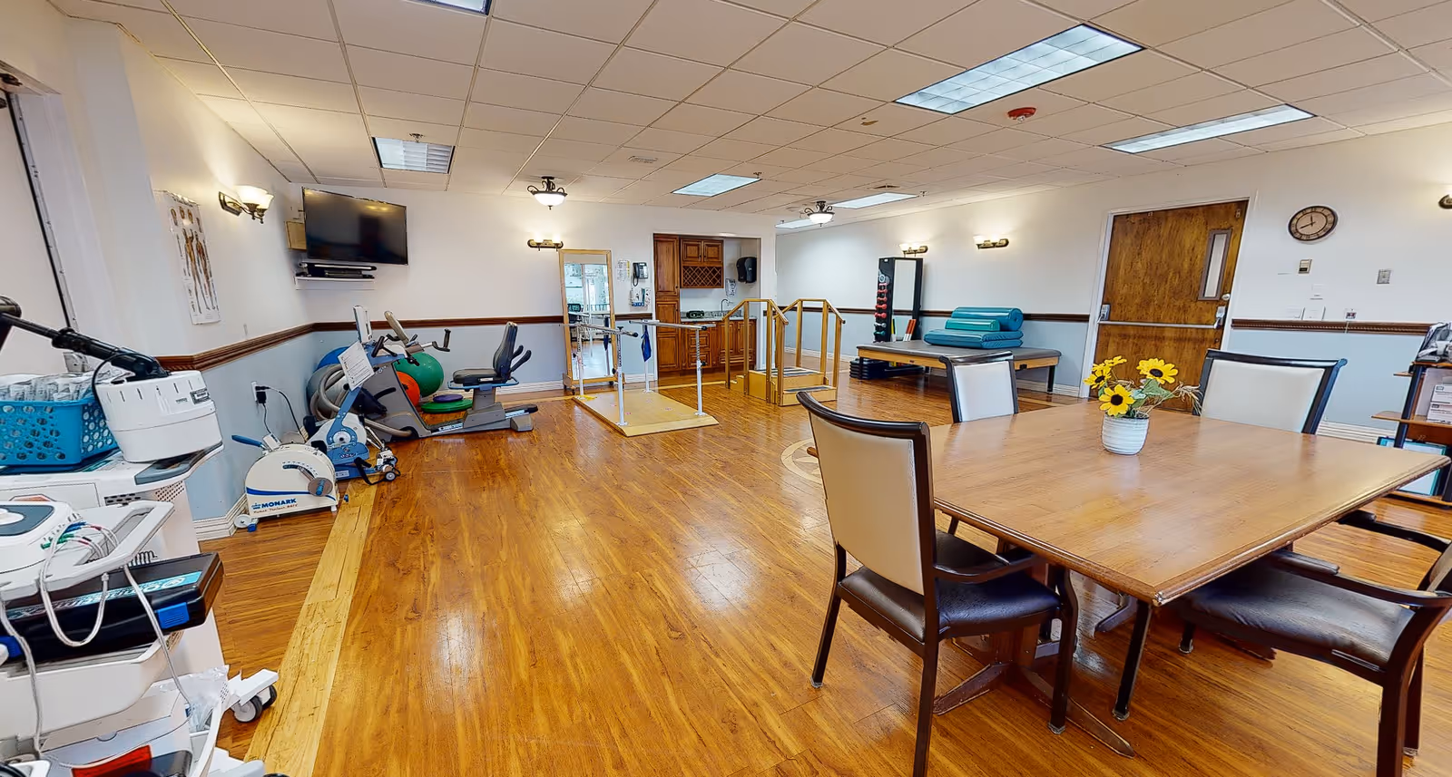 A spacious rehabilitation therapy room with exercise equipment, parallel bars, and a table with chairs.