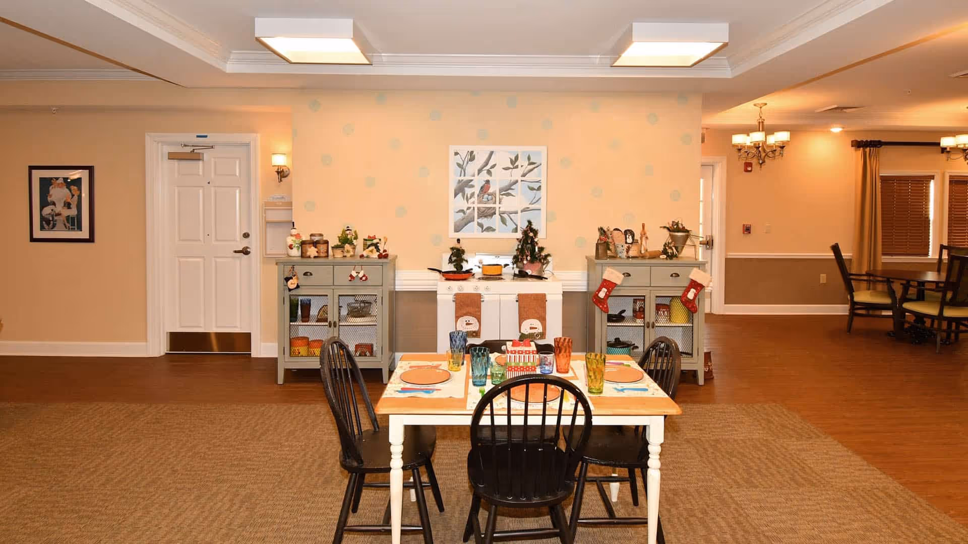 A cozy dining area in a senior living facility with a wooden table set for four, featuring colorful glasses and plates. Behind the table are two gray cabinets decorated with small holiday ornaments and stockings, and a white mantelpiece with more festive decorations. The walls are painted a soft beige with a framed bird artwork hanging above the mantel. The room has warm lighting and wooden flooring with a carpeted section under the table.