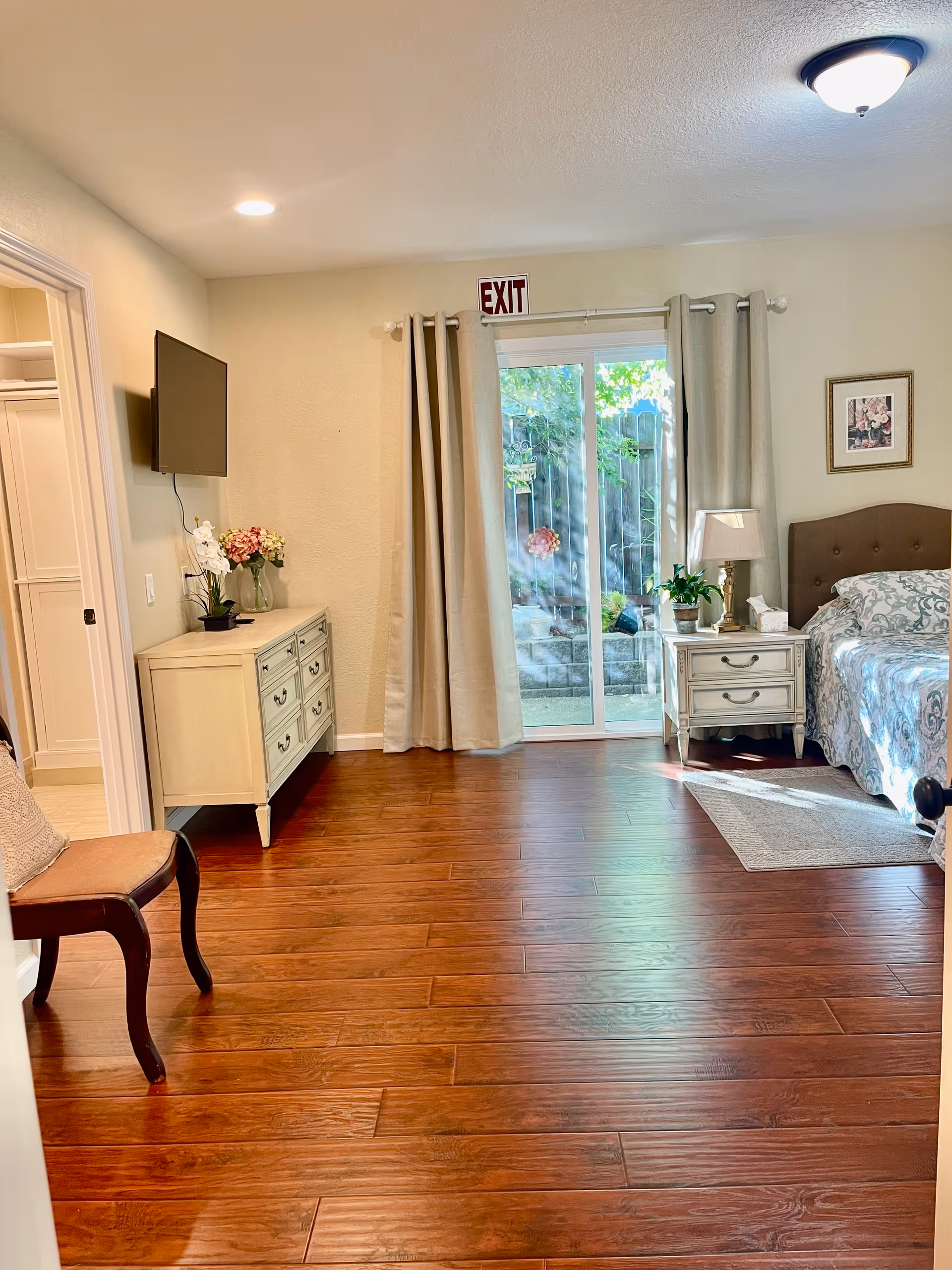 A bright bedroom with wooden flooring, a bed with patterned bedding, a nightstand with a lamp and plant, a dresser with flowers, a wall-mounted TV, a chair, and sliding glass doors with curtains leading to an outdoor area. An exit sign is visible above the sliding doors.