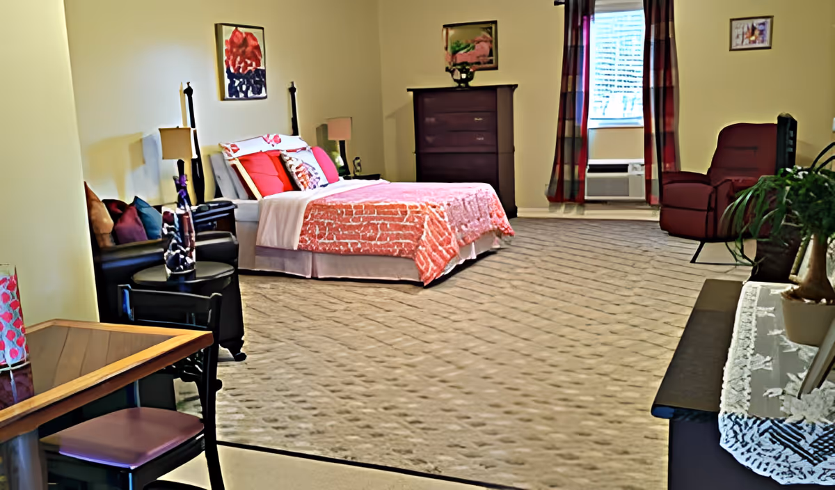 A spacious senior living bedroom with a bed covered in a red and white patterned bedspread, two bedside tables with lamps, a dark wooden dresser, a red armchair near a window with red and plaid curtains, a small table with a chair, and decorative artwork on the walls.