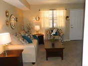 A cozy living room with a beige sofa adorned with blue and patterned cushions, a patterned armchair, a wooden coffee table with a small plant, a side table with a lamp, decorative wall mirrors, and a window with sheer curtains letting in natural light.