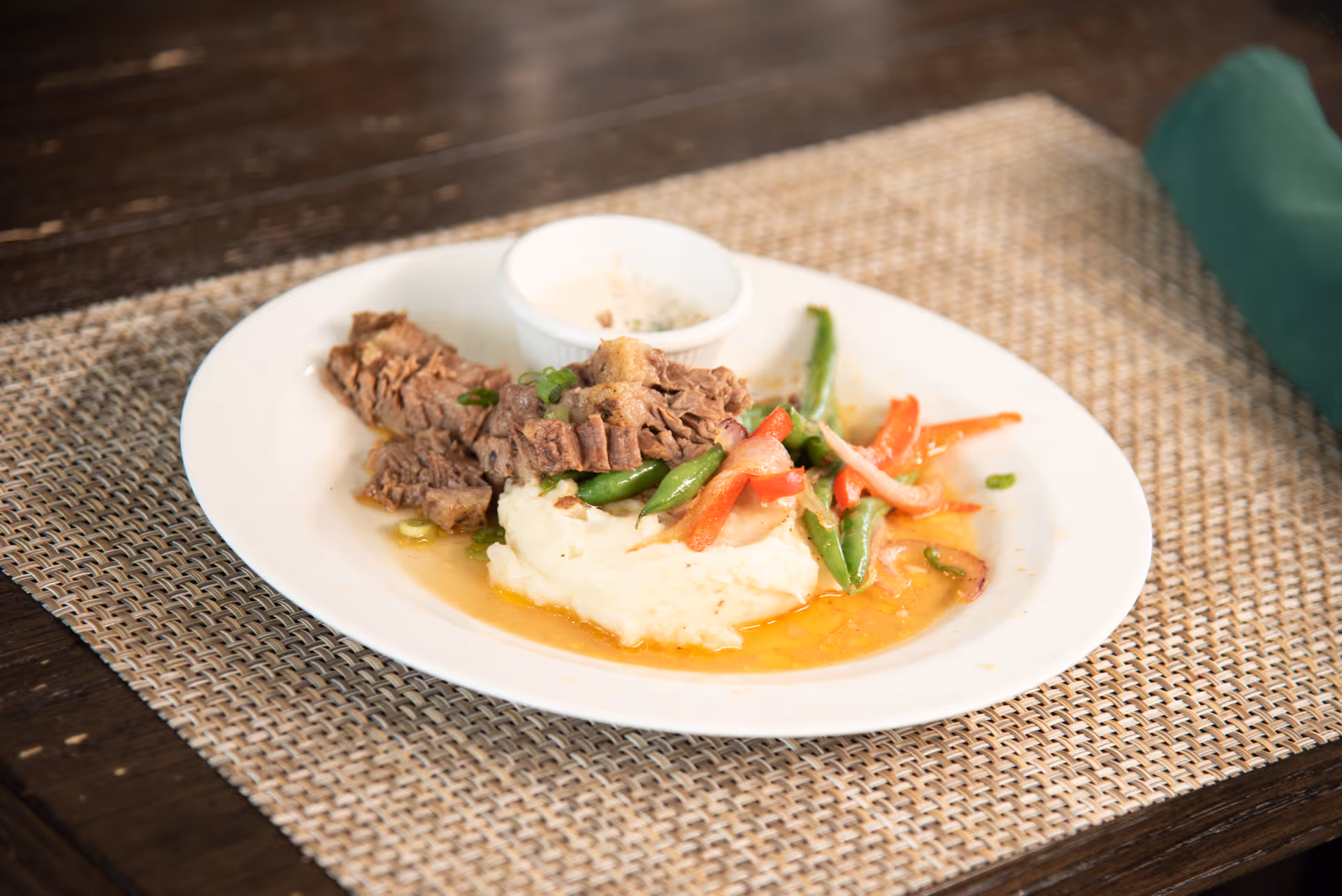 A white plate with a serving of sliced beef, mashed potatoes topped with green beans and red bell peppers, and a small cup of white sauce on the side, placed on a woven placemat on a wooden table.