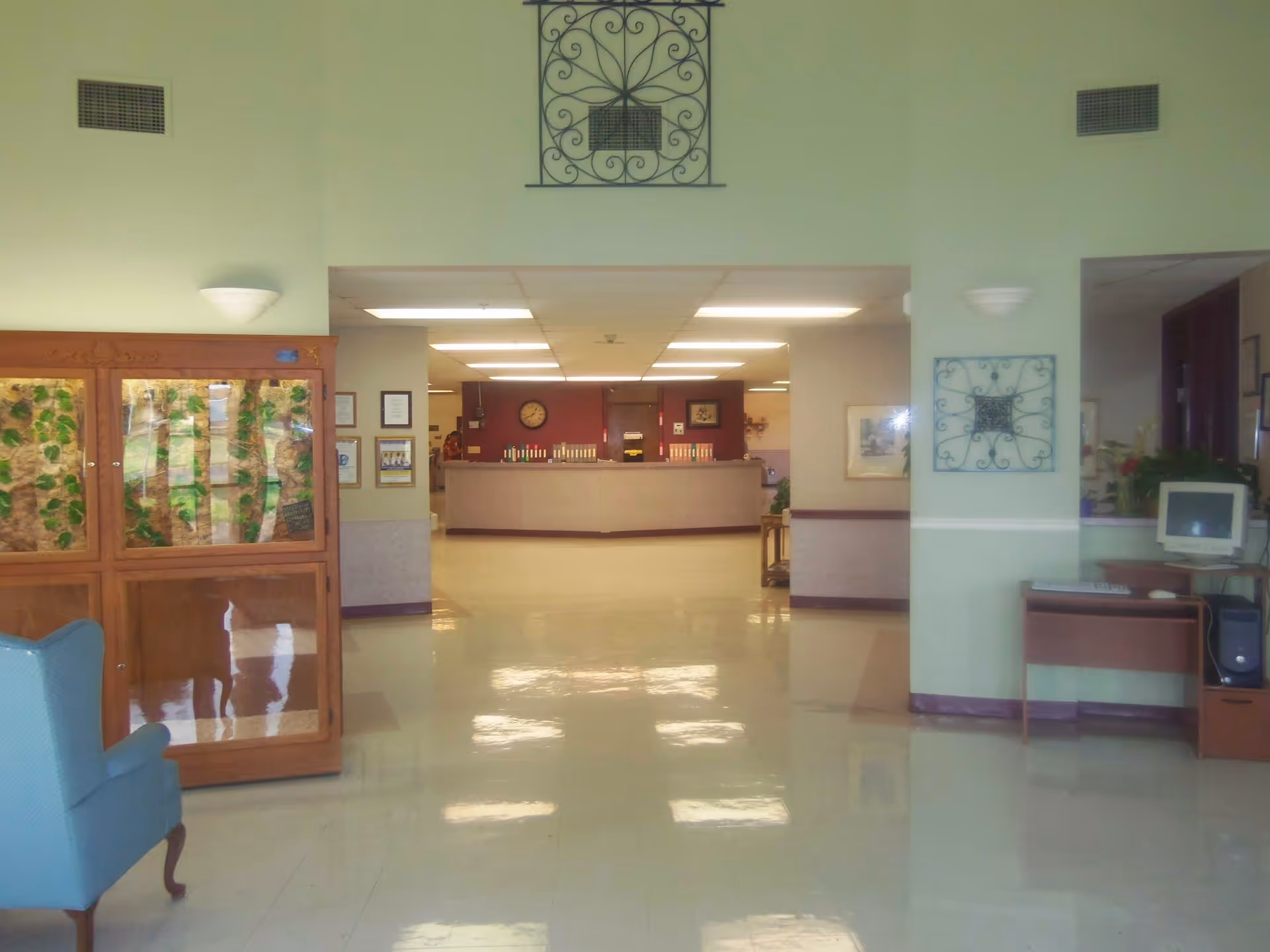 Interior view of a senior living facility lobby area with a reception desk in the background, a blue armchair on the left, a wooden display cabinet with glass doors, and a computer workstation on the right. The floor is shiny and reflective, and the walls are painted light green with decorative wall art.