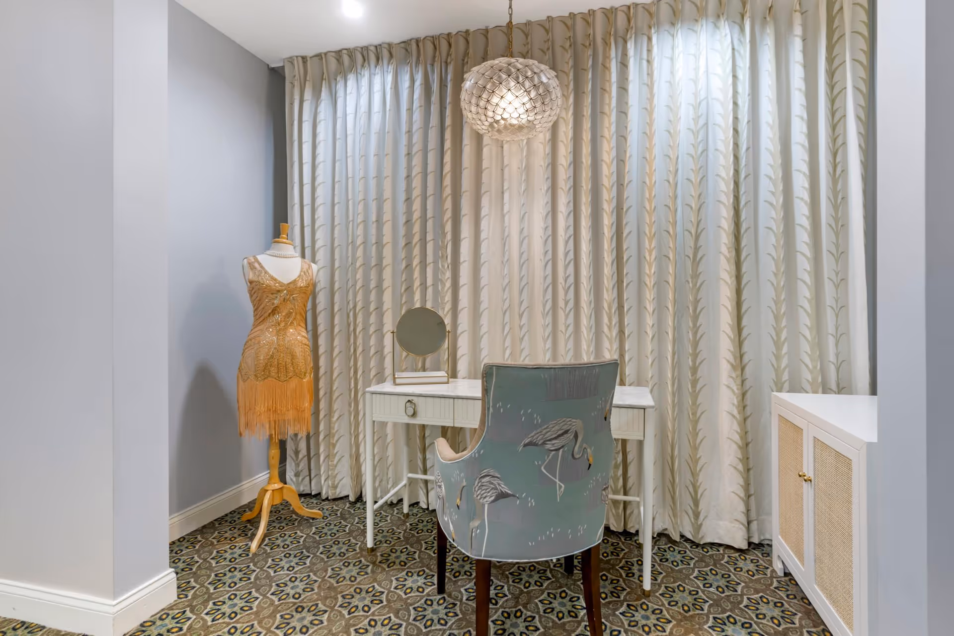 A small interior room with a patterned carpet, a white desk with a small round mirror on top, a chair with a bird design, a mannequin dressed in a gold fringed dress, and tall curtains with a leafy pattern covering the window. A decorative hanging light fixture is above the desk.