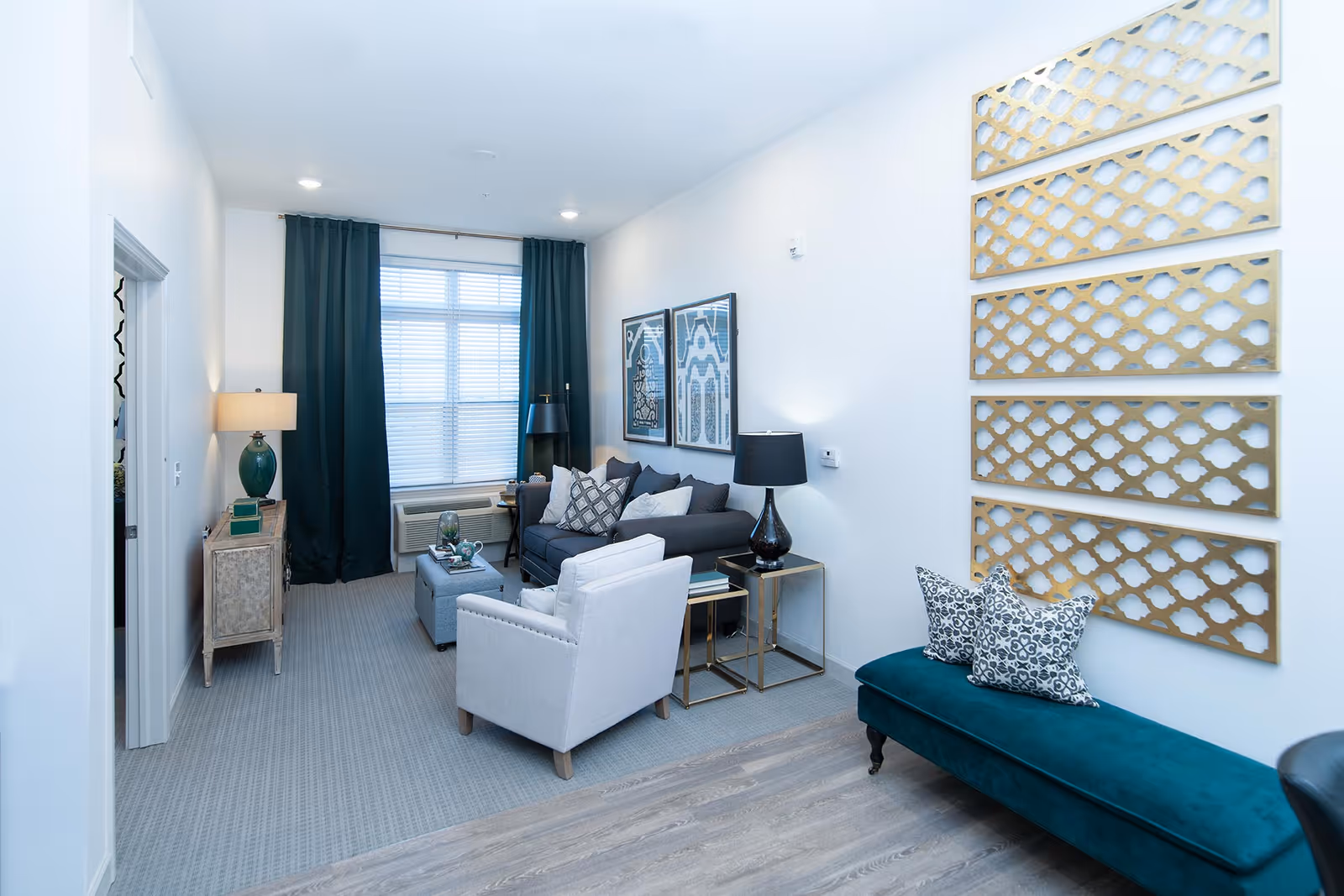 A modern living room with a gray sofa, a light gray armchair, and a teal upholstered bench with patterned pillows. The room features a window with dark curtains, two framed artworks on the wall above the sofa, a side table with a black lamp, a console table with a green lamp, and decorative gold wall panels on the right wall.