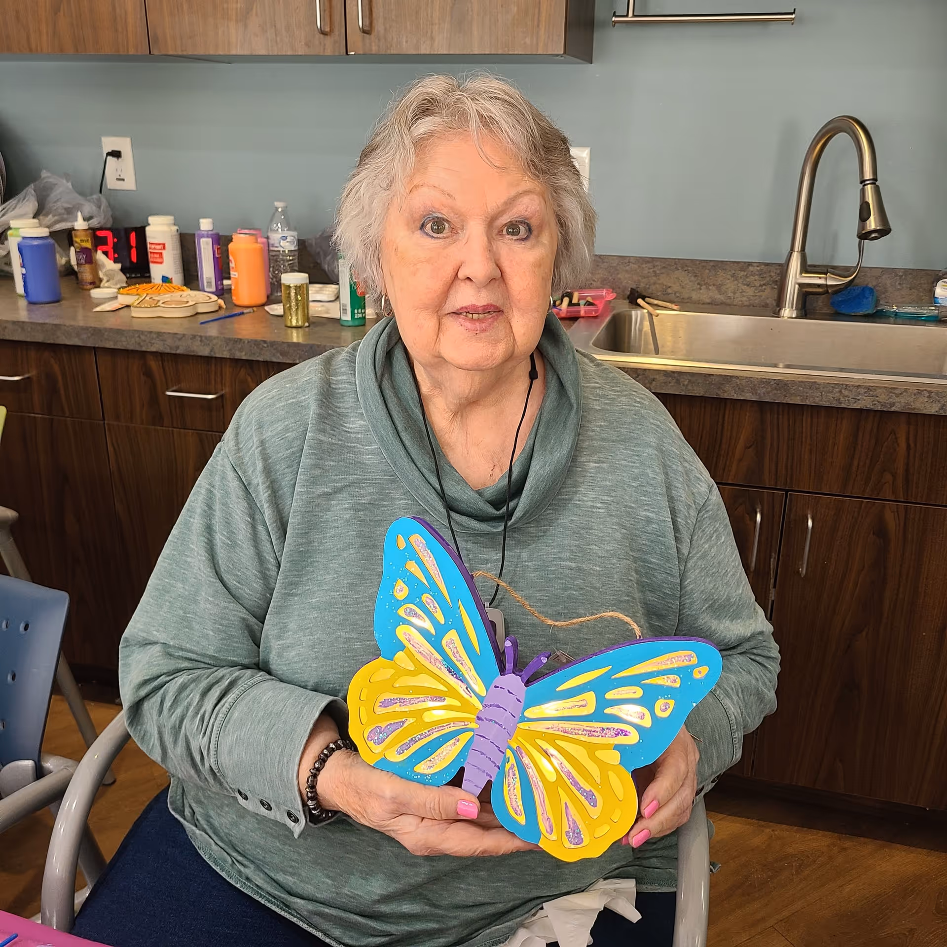 An elderly woman with gray hair wearing a green sweater is sitting in a chair in a kitchen-like room, holding a colorful wooden butterfly craft. Behind her are wooden cabinets, a countertop with various craft supplies, and a stainless steel sink with a faucet.